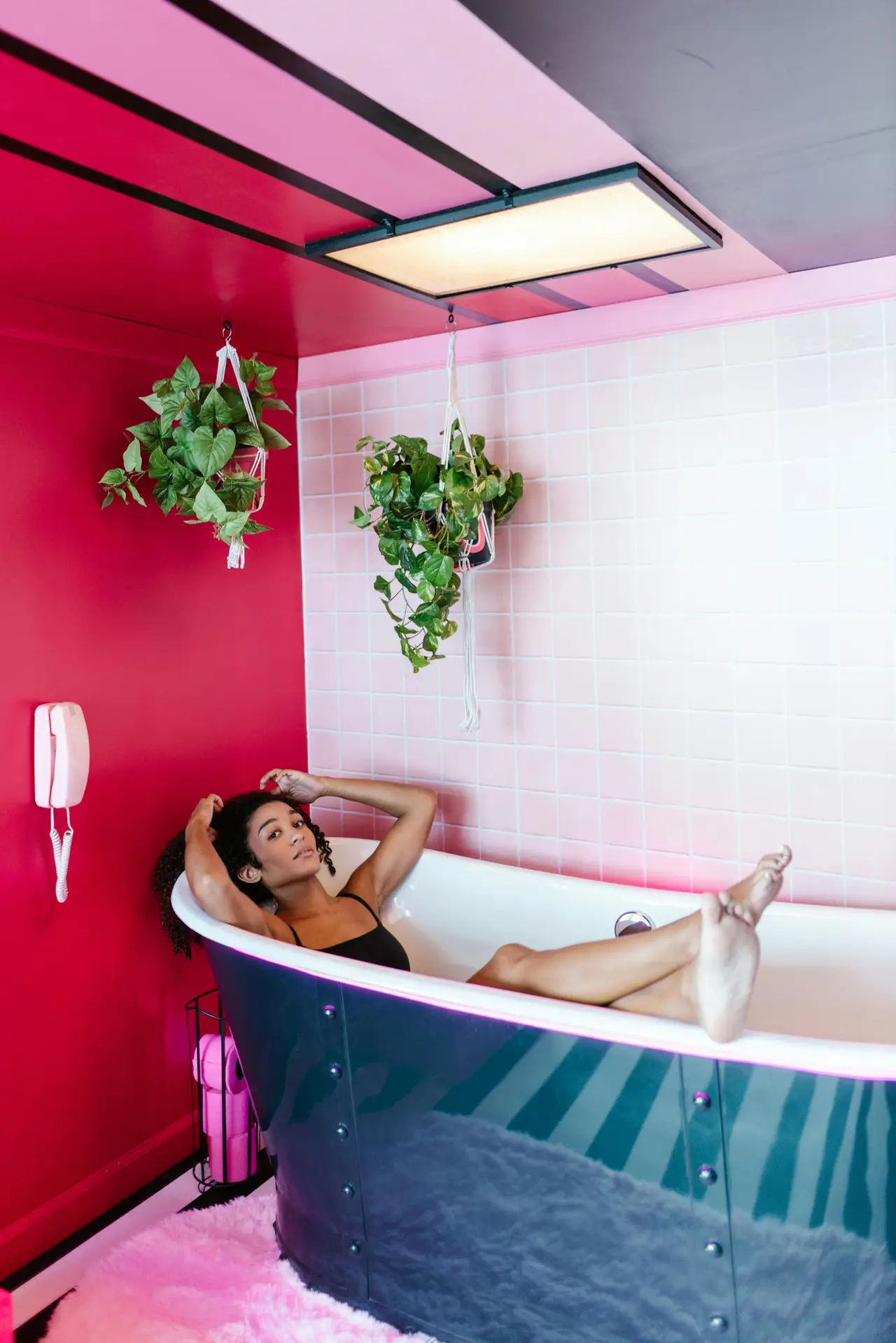A vibrant bathroom scene features a woman relaxing in a colorful tub with a glossy blue exterior, surrounded by pink walls, light tile accents, hanging plants, and modern lighting, presenting playful bathroom ideas with a tub that highlight how a colorful tub can turn bathing into a bold design statement.