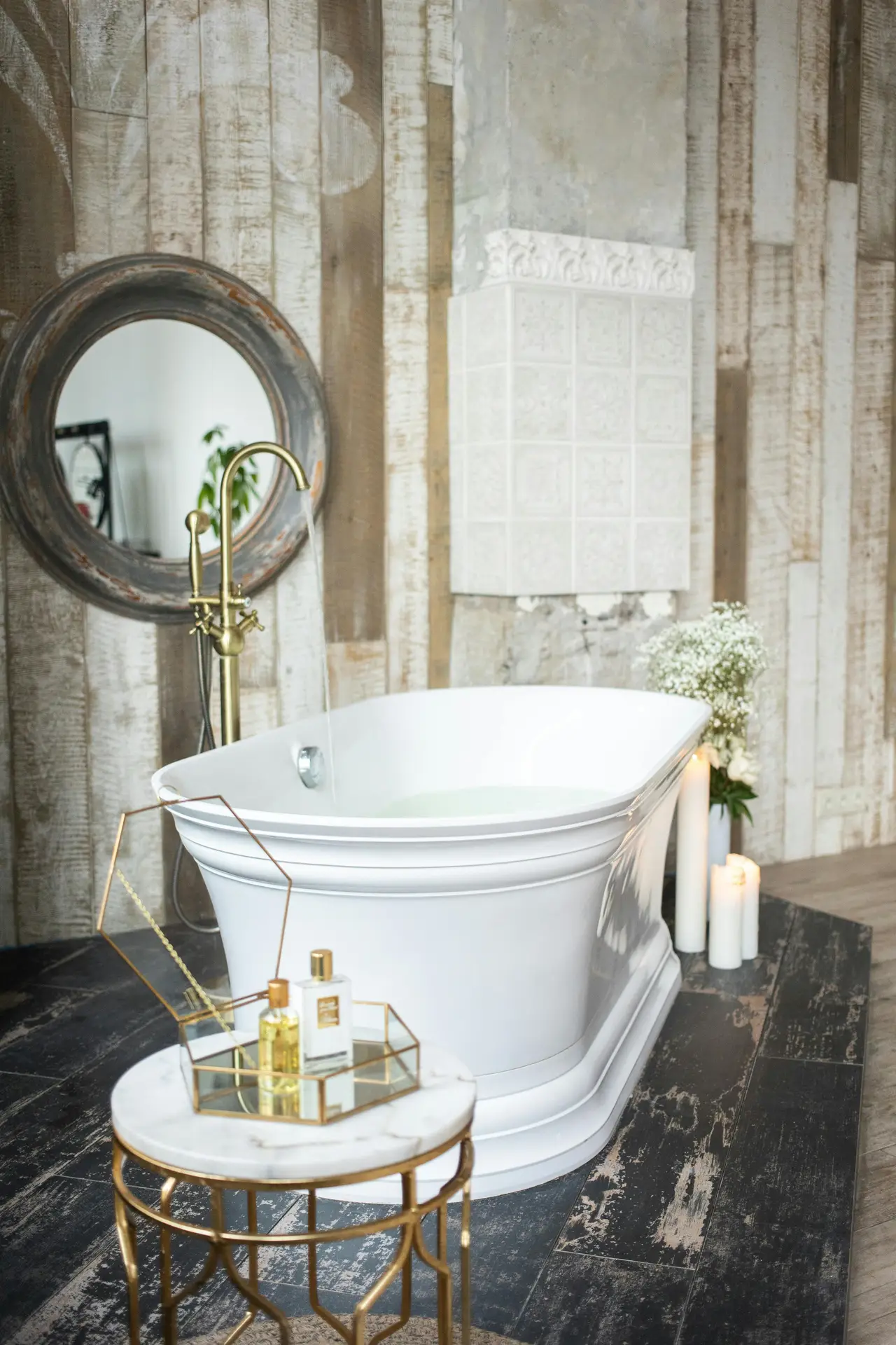 An elegant bathroom setup showcases a sculptural white bathtub with curved edges, paired with a vintage-style brass floor faucet, rustic wood-paneled walls, soft candlelight, and a small marble-topped side table with perfumes, creating luxurious bathroom ideas with a tub that feel made for kings and queens while still blending warmth and character.