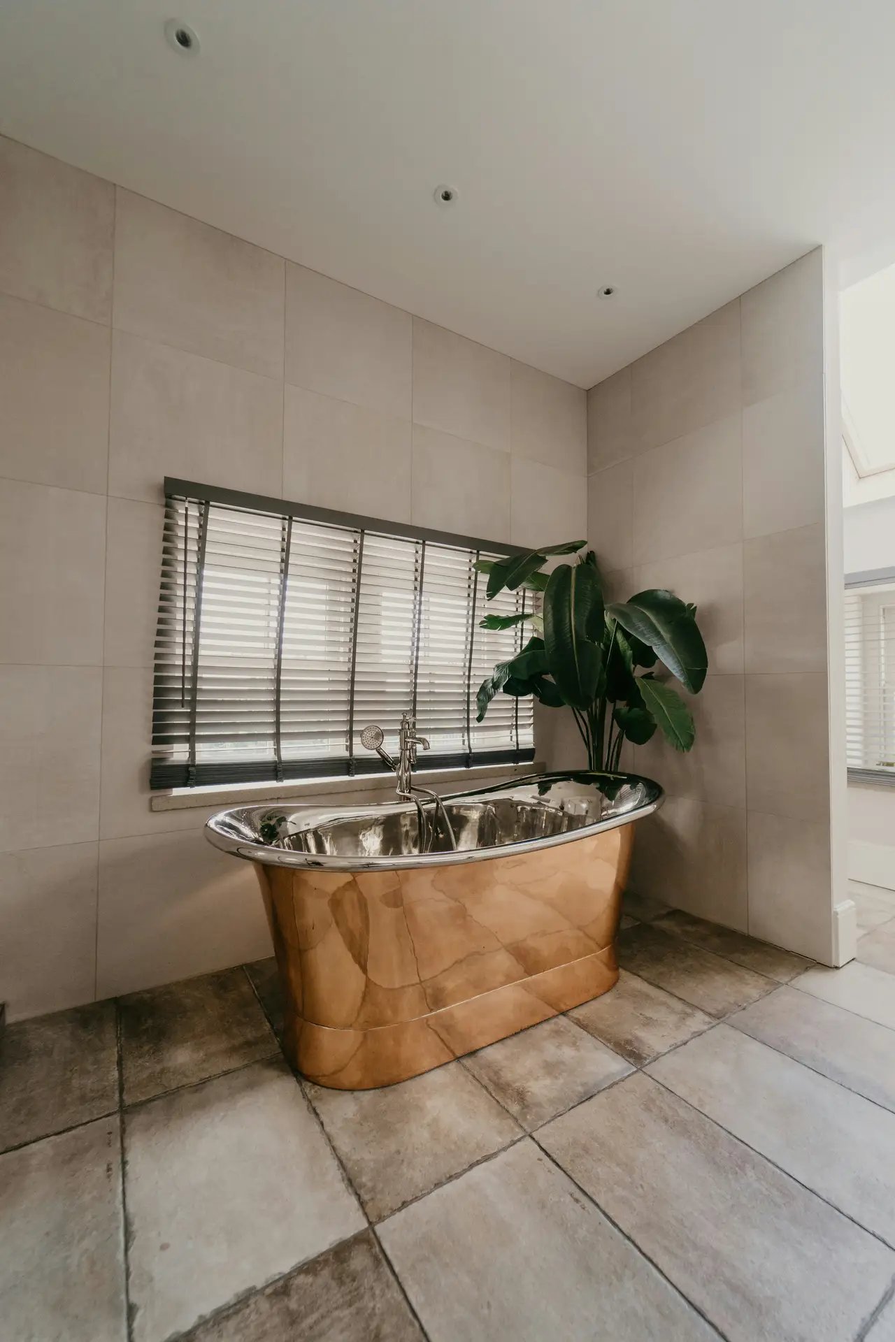 A minimalist bathroom interior showcases a sculptural metallic tub with a reflective finish, set against soft stone tiles, a wide window with blinds, and a leafy indoor plant, offering elegant bathroom ideas with a tub that blend modern materials, natural light, and a calm spa-inspired atmosphere.