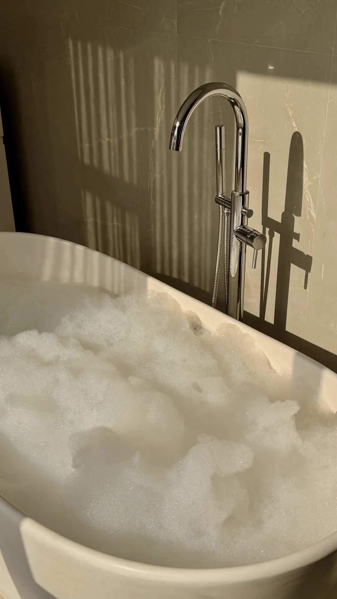 A freestanding minimalist tub filled with soft bubbles sits against a smooth stone wall, paired with a sleek chrome floor-mounted faucet and warm natural light, showcasing calm bathroom ideas with a tub that focus on simplicity, texture, and a spa-like atmosphere.
