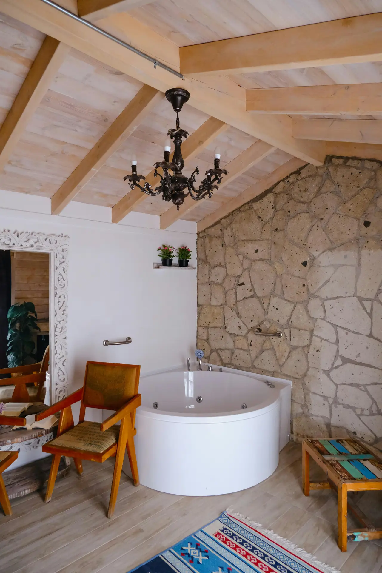 A bright, rustic-inspired bathroom features a round white soaking tub set against a textured stone wall, with exposed wooden ceiling beams, a vintage chandelier, wooden chairs, and decorative accents, offering relaxed bathroom ideas with a tub and showcasing a round tub as a cozy focal point.