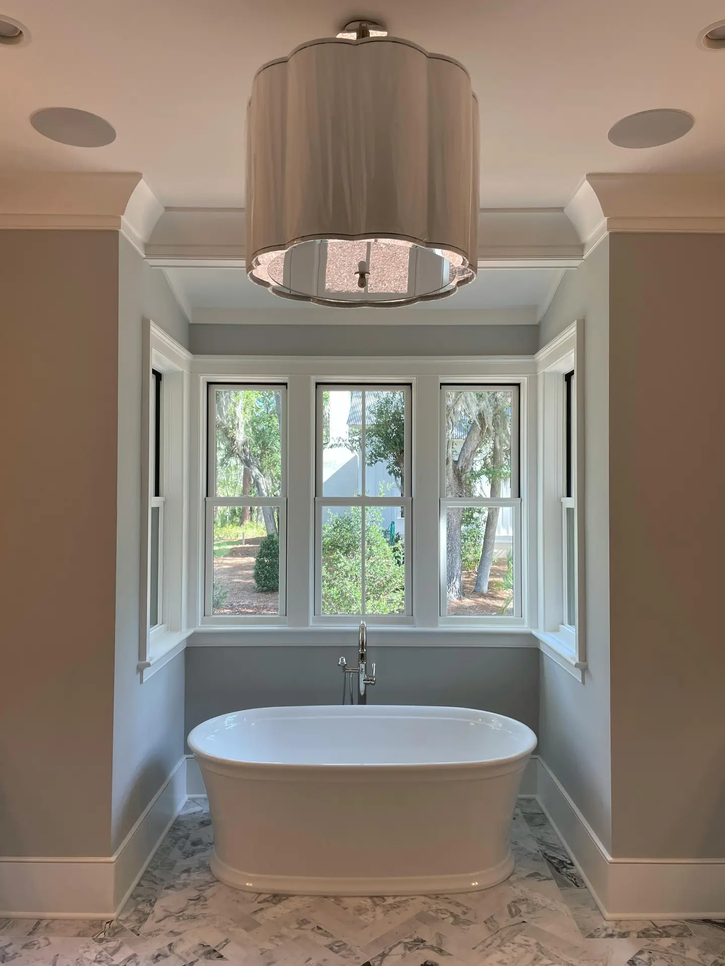A serene bathroom shows a freestanding white bathtub perfectly centered in a built-in alcove, framed by soft gray walls, a marble floor, and three tall windows with garden views, offering elegant bathroom ideas with a tub that highlight a tub in a niche as a calm, spa-like focal point.