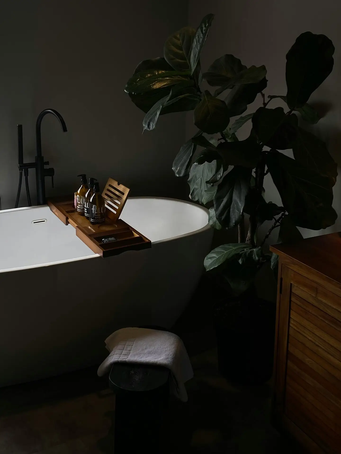 A sleek white freestanding bathtub sits against a dark, moody wall, styled with a wooden bathtub tray holding bottles and bath essentials, alongside a tall indoor plant and warm wood furniture, illustrating bathroom ideas with a tub and a tub with bathtub shelf that create a calm, spa-like atmosphere.