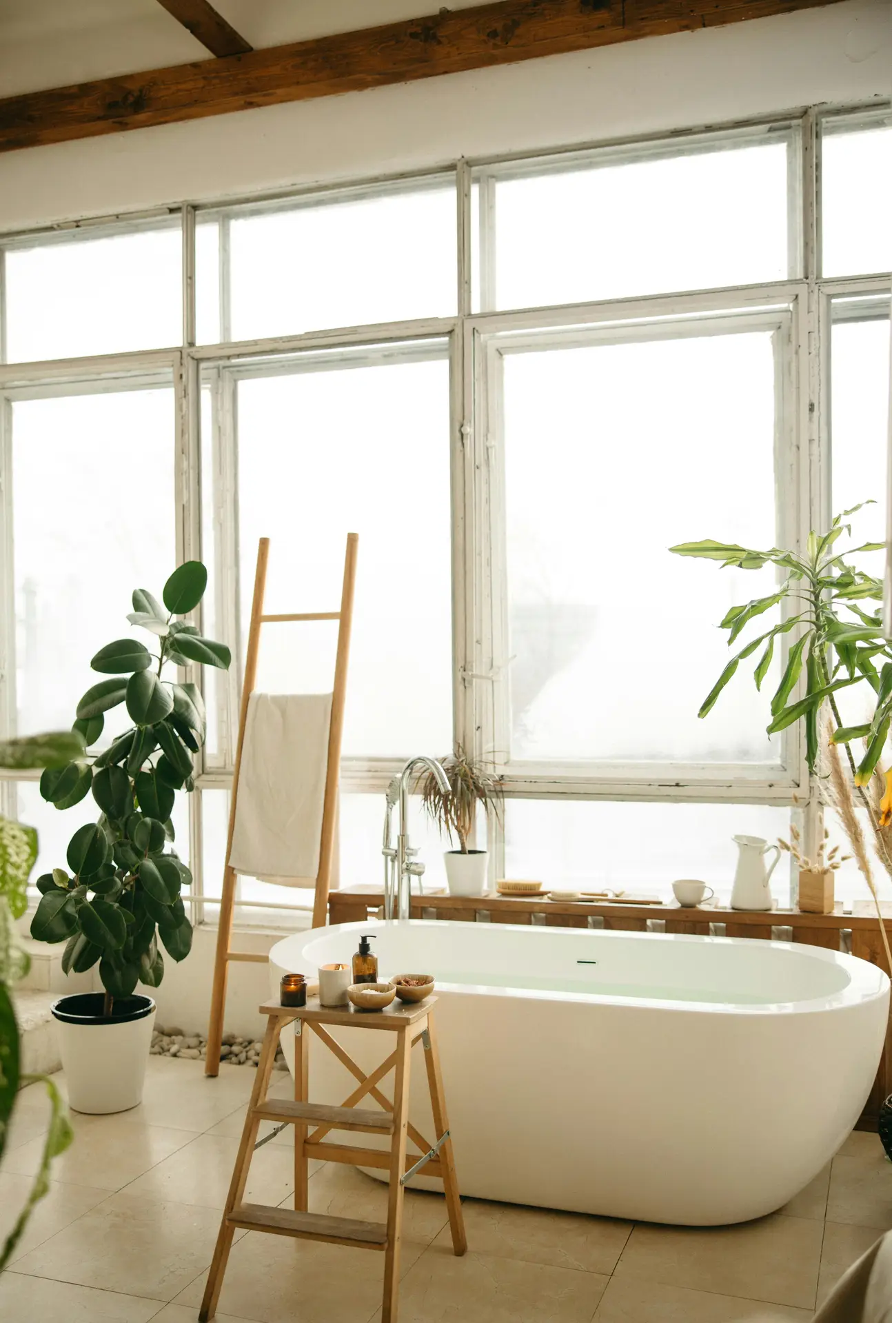 A light-filled bathroom showcases a modern white soaking tub with a wide rim placed in front of large industrial-style windows, surrounded by lush indoor plants, a wooden towel ladder, and a small stool with bath essentials, offering relaxed bathroom ideas with a tub and highlighting a tub with wide rim as a practical design feature.