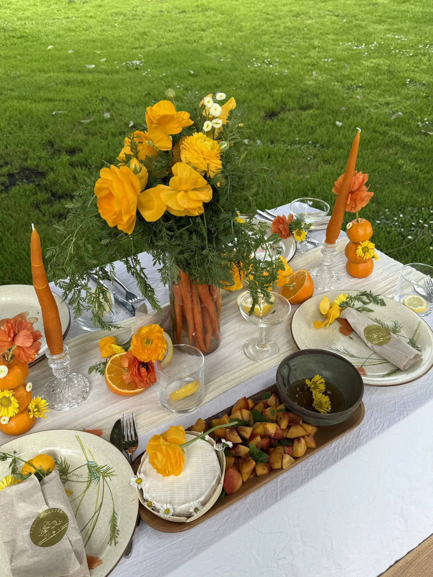 Bright easter table decor styled in an all yellow and orange color palette, featuring a carrot bouquet centerpiece, citrus accents, orange candles, fresh flowers, layered place settings, and shared dishes arranged on a long outdoor table set against a green lawn.