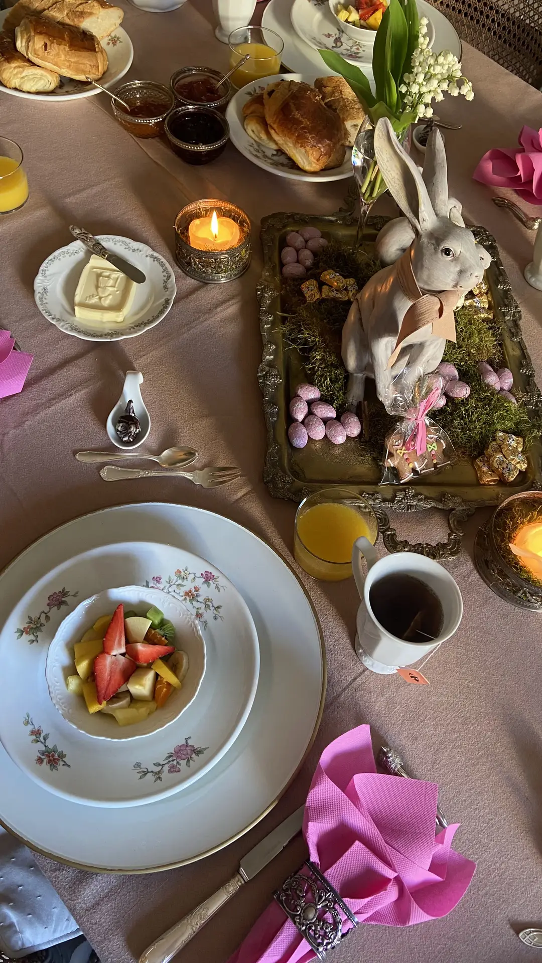 Easter table decor featuring a charming bunny centerpiece placed on a decorative tray with moss, pastel eggs, and small treats, surrounded by a beautifully set breakfast table with croissants, fruit bowls, candles, tea cups, vintage plates, and soft pink napkins, creating a warm and inviting holiday atmosphere.