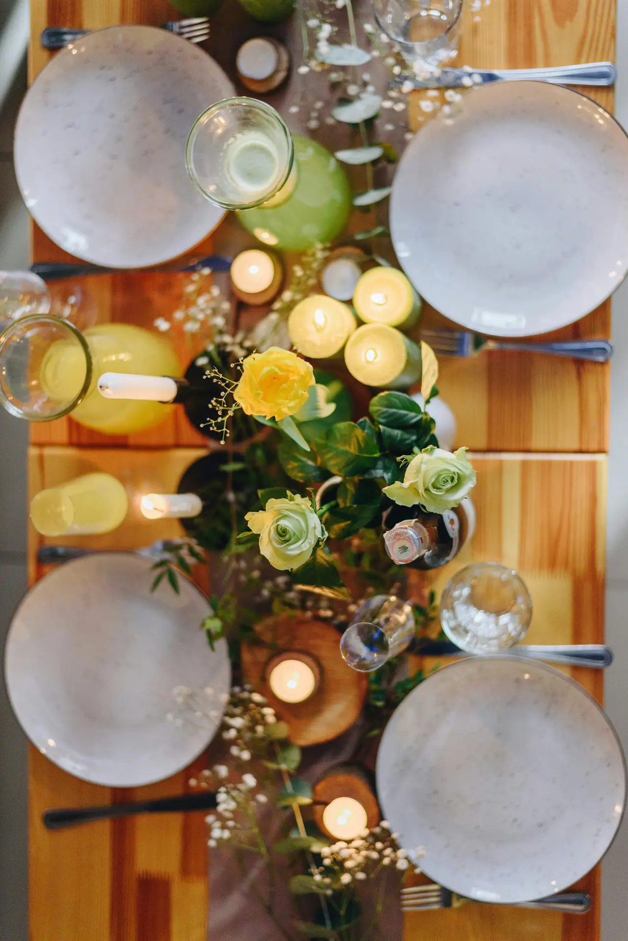 Overhead view of elegant easter table decor featuring candles, yellow roses and baby's breath arranged along a wooden table, with neutral ceramic plates, glassware, soft greenery, and warm candlelight creating a cozy yet refined spring table setting.