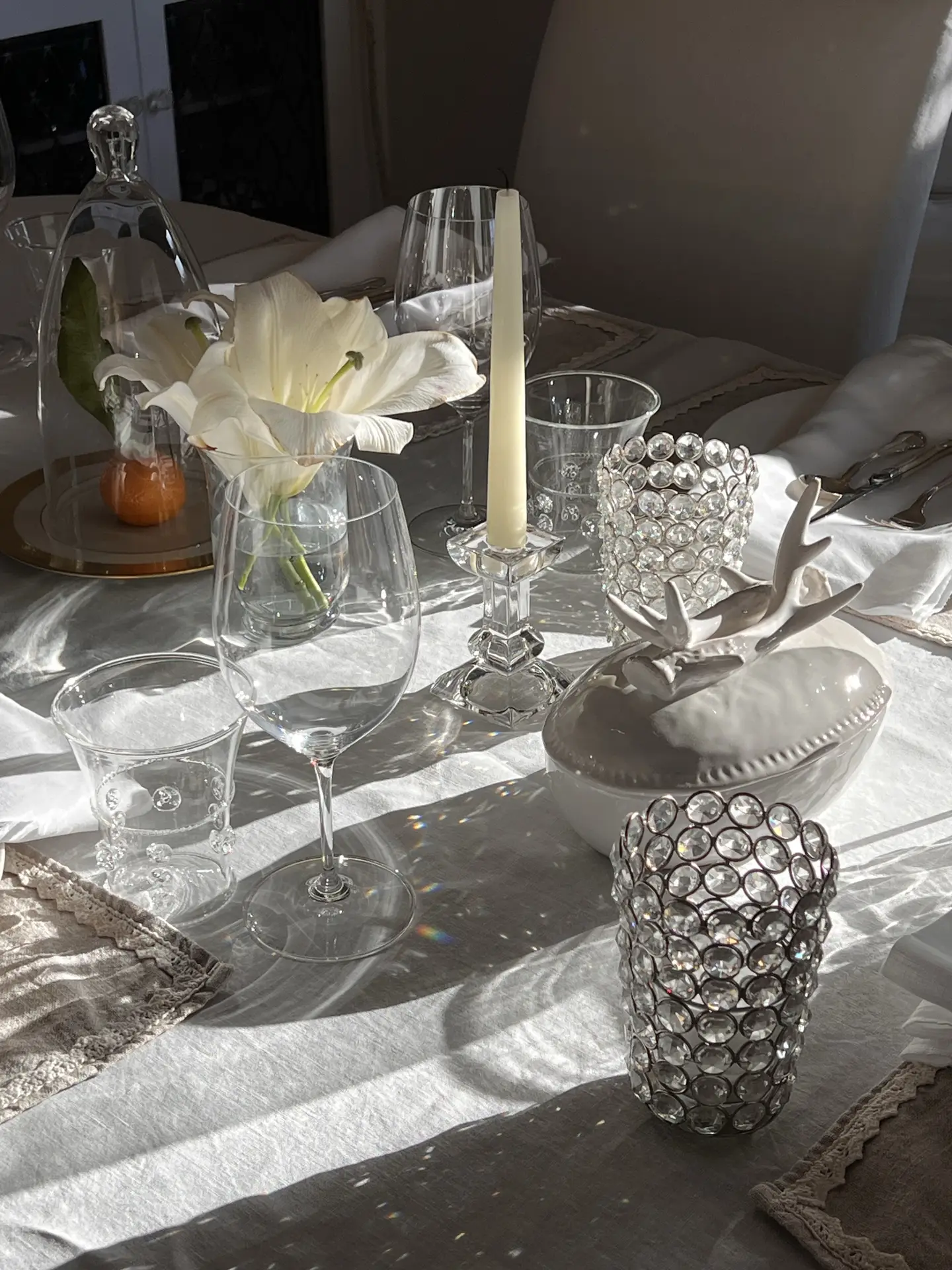 Elegant easter table decor styled with decent decor elements, featuring a soft white tablecloth, clear glassware, a single white taper candle in a crystal holder, delicate floral accents in small vases, and subtle ceramic details that create a light, refined, and inviting holiday table setting.