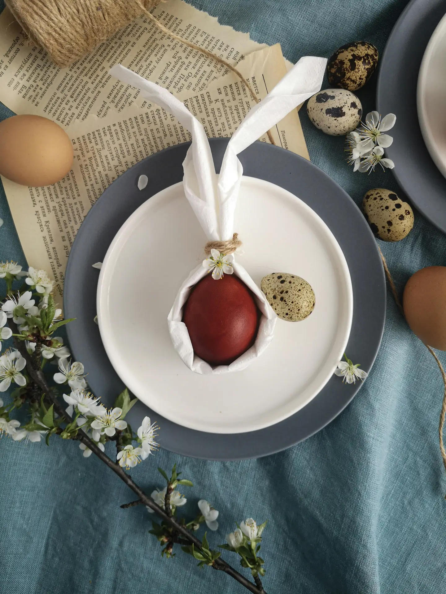 Flat lay of easter table decor showing an easter bunny egg decor variation, with a red dyed egg wrapped in a white napkin shaped like bunny ears on layered plates, surrounded by speckled eggs, delicate spring blossoms, vintage book pages, and a soft blue linen tablecloth for a natural seasonal look.