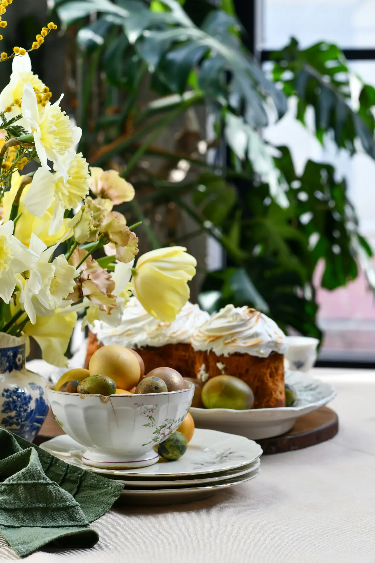 Easter table decor styled with eggs in a bowl and flowers, showing a vintage-style ceramic bowl filled with naturally dyed eggs, fresh yellow spring blooms in a patterned vase, layered plates, a soft linen tablecloth, and a frosted cake in the background, creating a relaxed and elegant seasonal table setting.