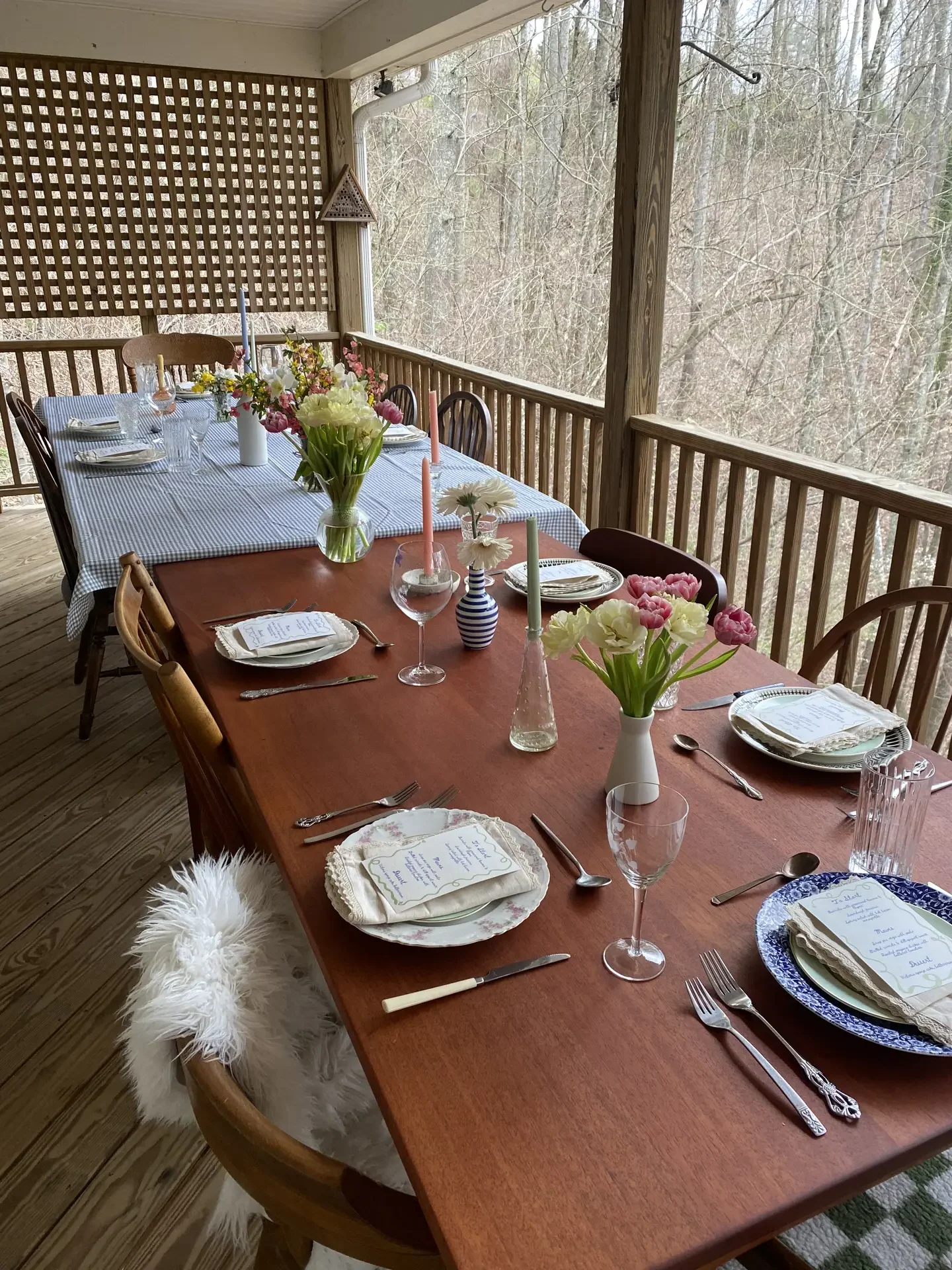 Bright easter table decor set on a covered porch, featuring multiple vases with white, pink and yellow flowers arranged along a wooden dining table, paired with layered plates, handwritten place cards, pastel candles, and classic glassware, creating a welcoming spring atmosphere surrounded by natural outdoor views.