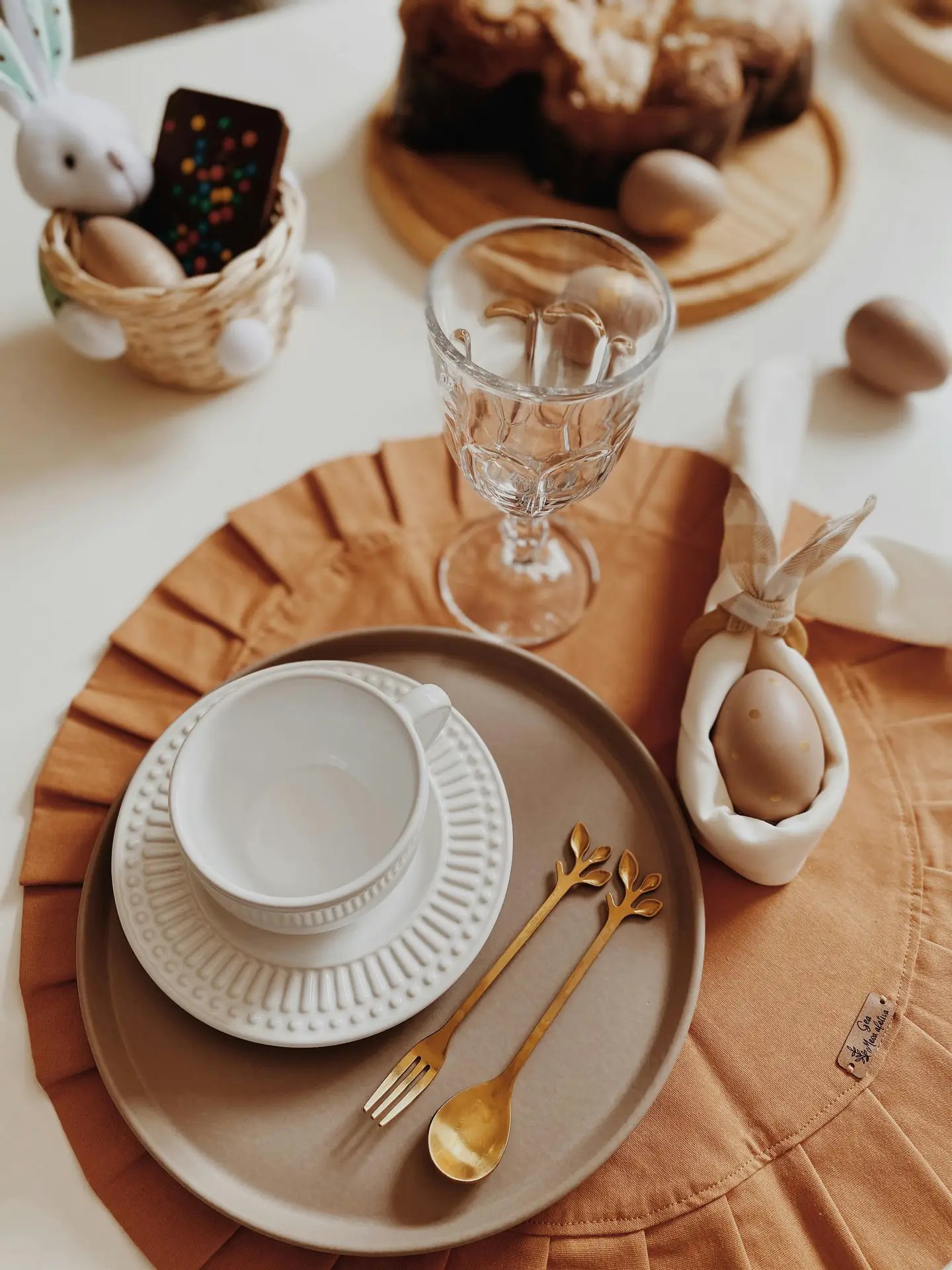 Warm and inviting easter table decor with natural decor with bunny details, featuring layered neutral plates, a white cup, gold-toned cutlery, a soft fabric napkin holding a speckled egg, and subtle bunny accents alongside rustic treats and glassware for a cozy spring-inspired setting.