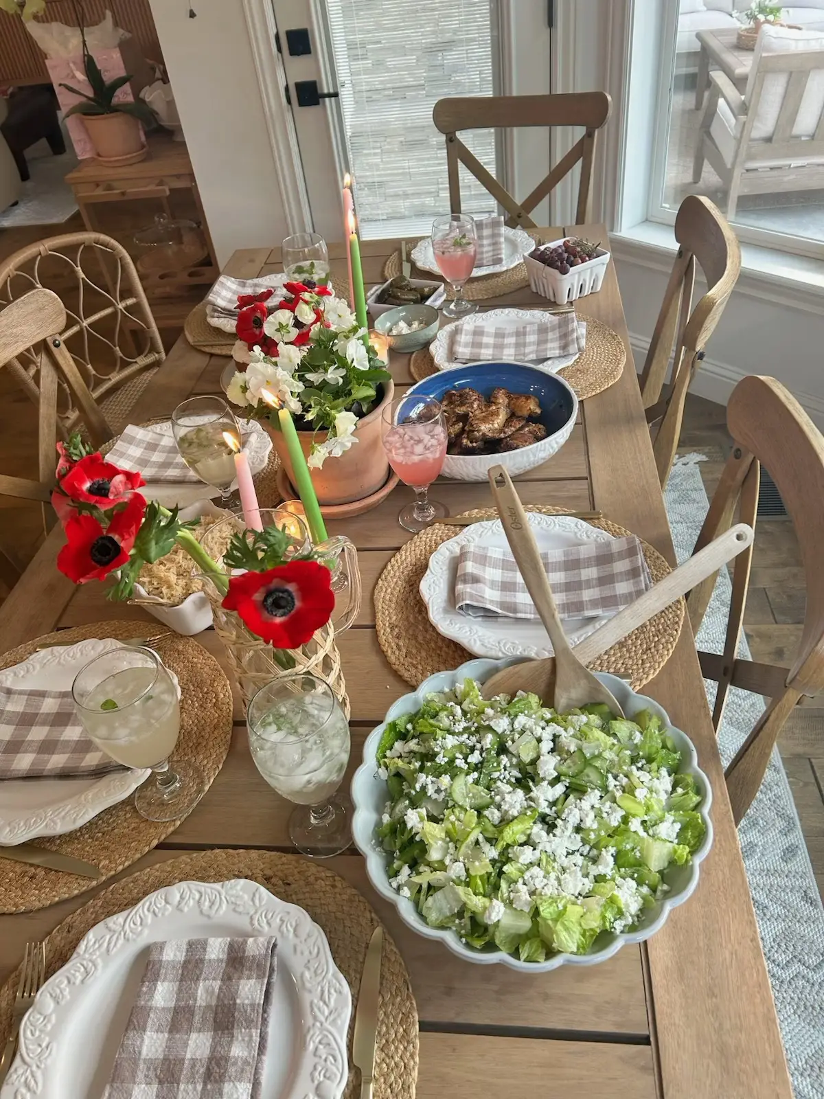 Easter table decor featuring poppy and white flowers arranged in simple vases along a wooden dining table, paired with woven placemats, folded neutral napkins, pastel candles, glassware, and shared serving dishes that create a warm and inviting spring dining atmosphere.