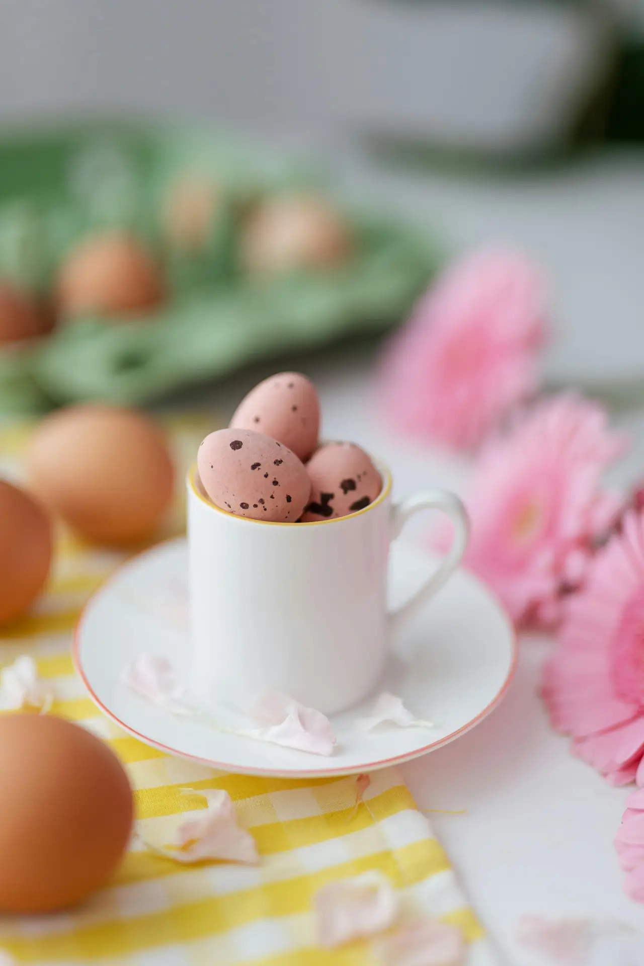 Soft and playful easter table decor showing small eggs in cup placed on a white saucer, surrounded by pastel flower petals, fresh pink blooms, and a yellow gingham cloth that adds a light and cheerful spring mood.
