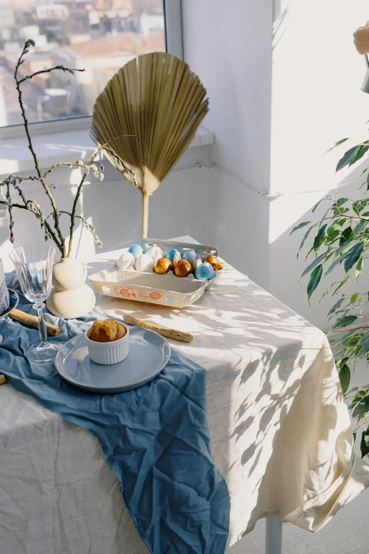 Bright and airy easter table decor featuring a soft table runner and colorful eggs arranged in a rustic carton, paired with simple tableware, natural branches in vases, and warm sunlight creating a calm spring atmosphere near the window.