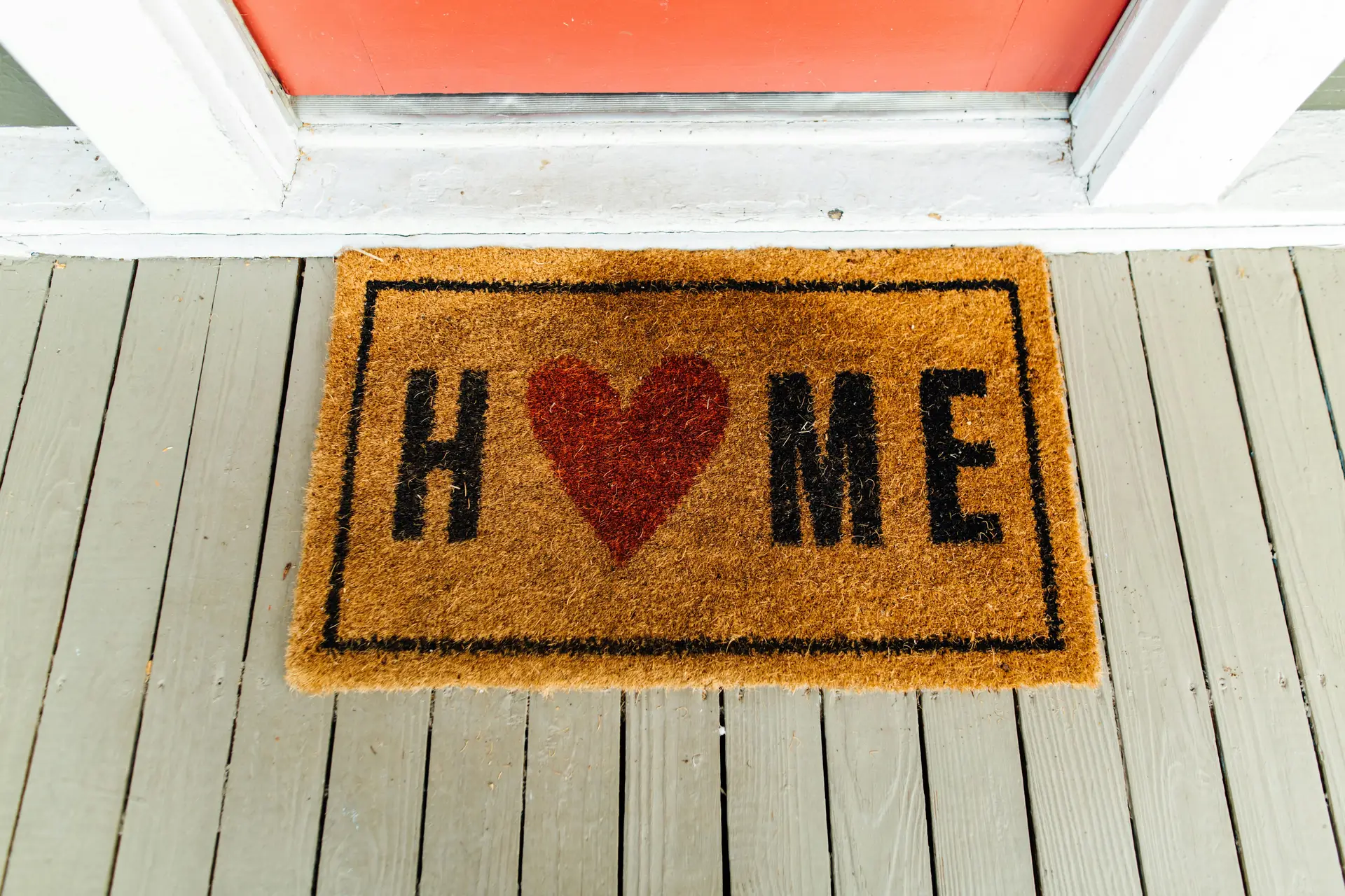 Front porch decor ideas for spring featuring a natural fiber door mat with a heart motif and the word “home,” placed on a light wooden porch floor in front of a brightly painted entry door for a warm and welcoming first impression.