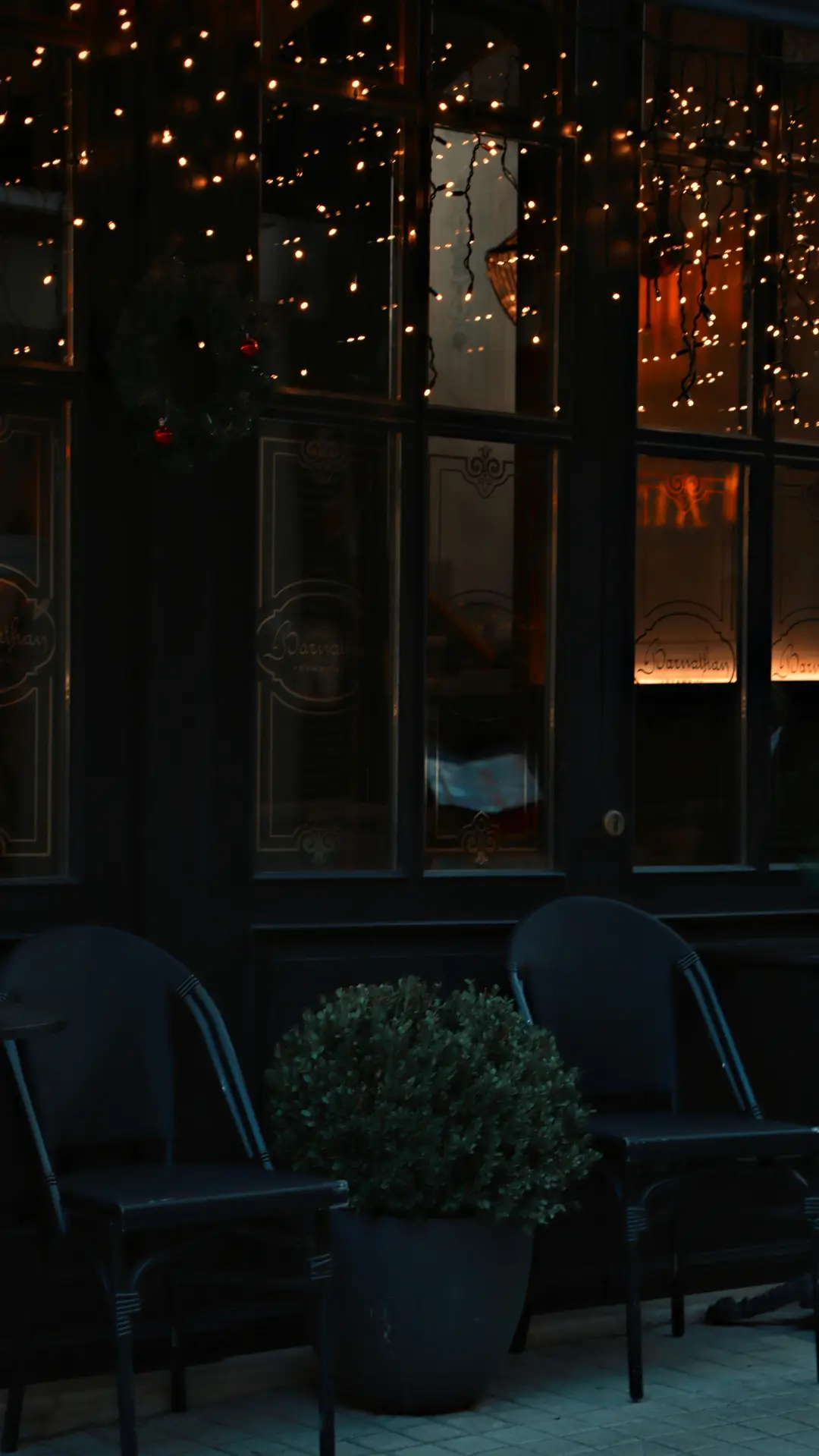 Front porch decor ideas for spring showcasing a moody evening porch with warm fairy Lights draped across dark window panels, black outdoor chairs lined up neatly, and a simple potted shrub adding a soft, cozy glow to the entrance after sunset.