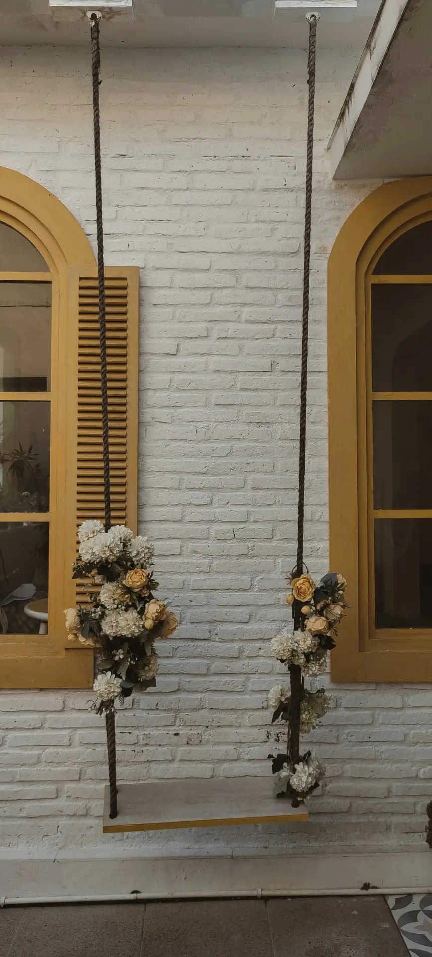 Front porch decor ideas for spring showcasing a flower-decorated swing suspended by rustic ropes, set against a white brick wall with warm yellow shutters, blending soft floral accents with a charming, romantic porch design.