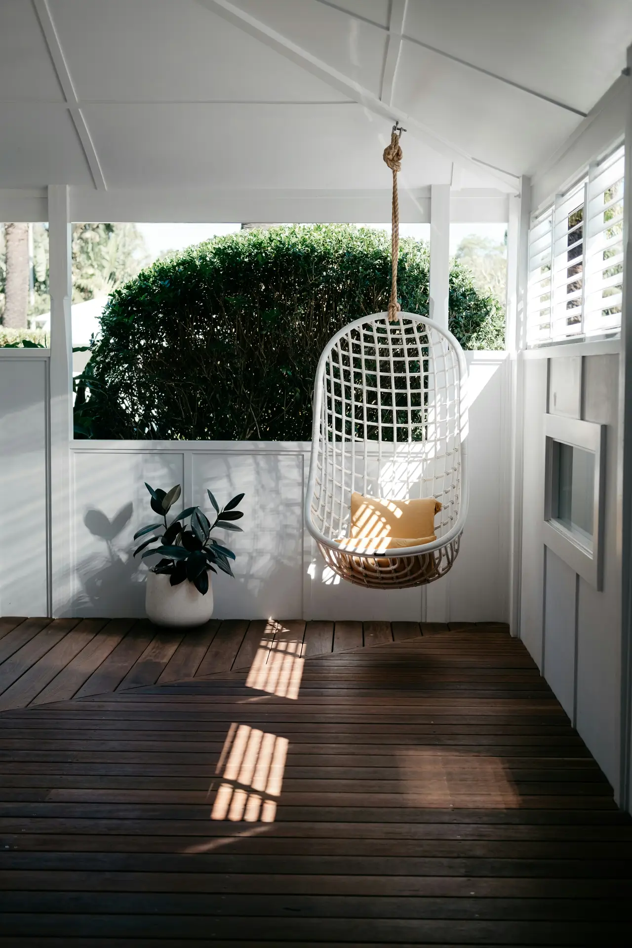 Front porch decor ideas for spring featuring a hanging chair suspended from the ceiling, paired with warm wood flooring, white panel walls, and a simple potted plant that creates a calm, airy outdoor seating nook.