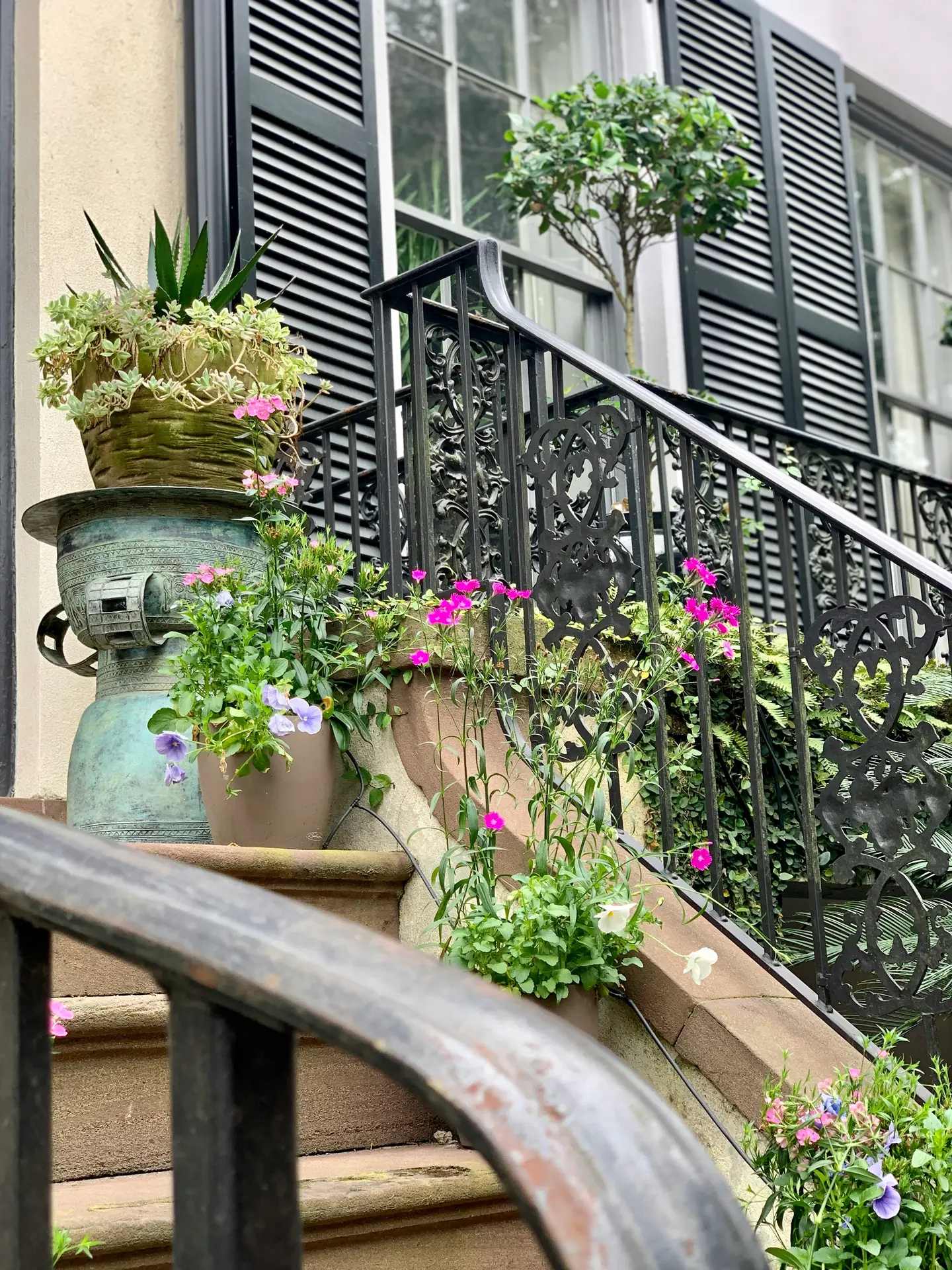 Front porch decor ideas for spring showcasing stone steps framed by ornate black railings and large planters filled with layered greenery, succulents, and bright seasonal flowers, creating a lush and welcoming entrance with a classic townhouse feel.