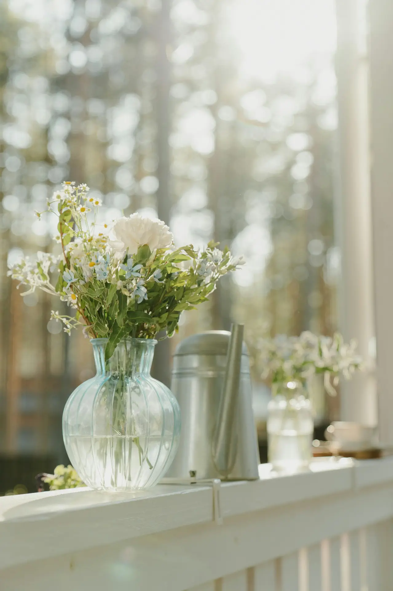 Front porch decor ideas for spring featuring a bright railing with flower vases, where delicate wildflowers and greenery sit in clear glass jars alongside a metal watering can, softly lit by natural sunlight and framed by a blurred woodland backdrop for a fresh, relaxed seasonal look.