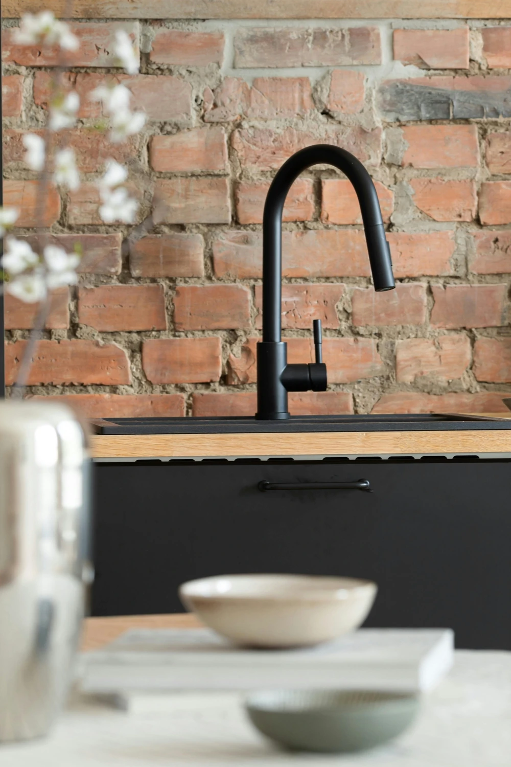 Modern kitchen ideas featuring matte black fixtures, with a sleek black faucet mounted on a wooden countertop, dark cabinetry below, and a warm exposed brick backsplash, creating a balanced mix of industrial and contemporary design with soft, neutral tableware in the foreground.