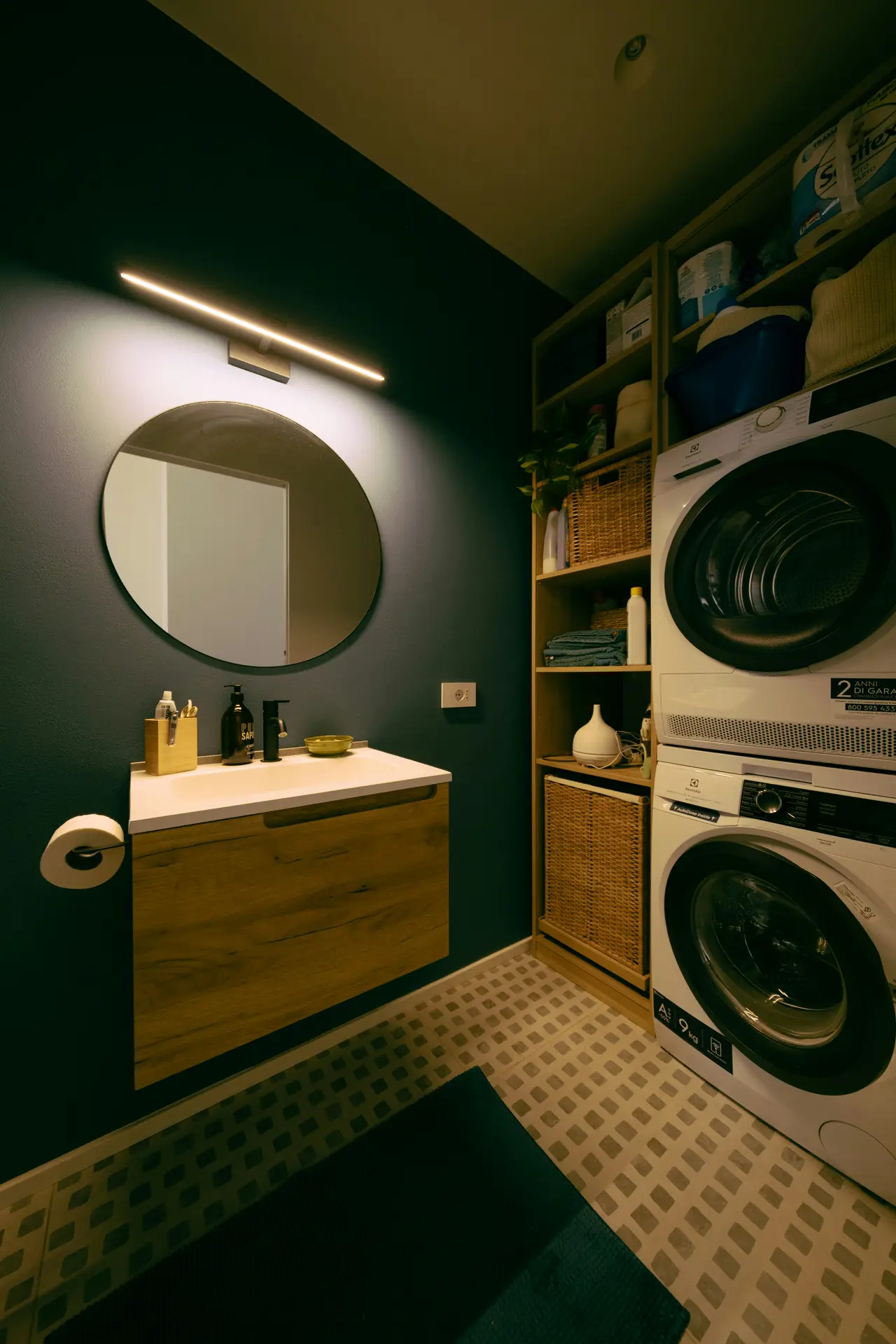 A stylish, compact laundry space with a floor-to-ceiling laundry wall featuring stacked washer and dryer, open shelving with baskets and supplies, and a small vanity with mirror and lighting, illustrating laundry room organization ideas that blend storage, function, and a polished, spa-like look.