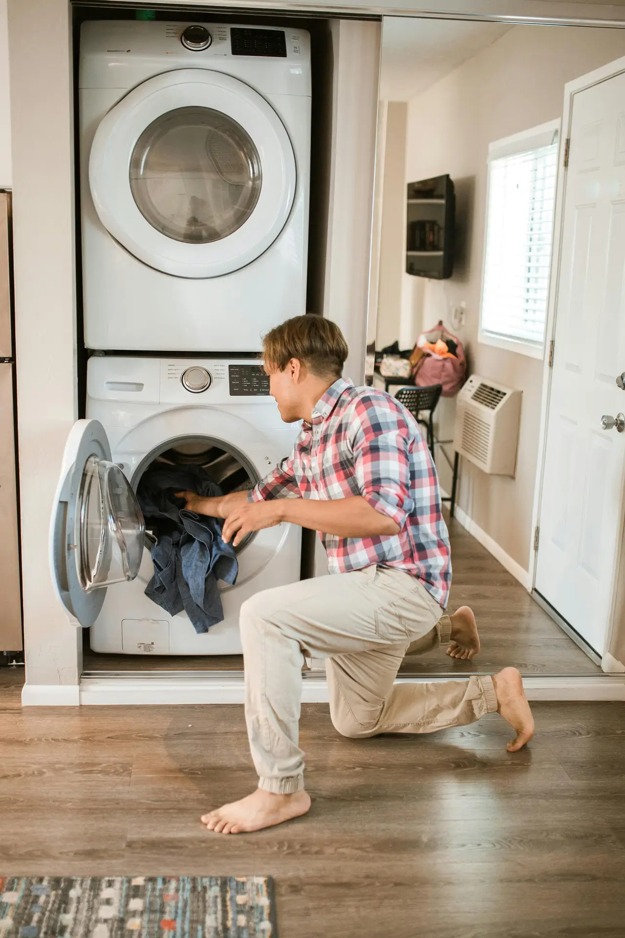 A person kneels in front of a stacked washer and dryer tucked neatly into an integrated laundry niche, loading clothes while the appliances blend into the surrounding living space, illustrating laundry room organization ideas that make everyday tasks functional and unobtrusive in modern homes.