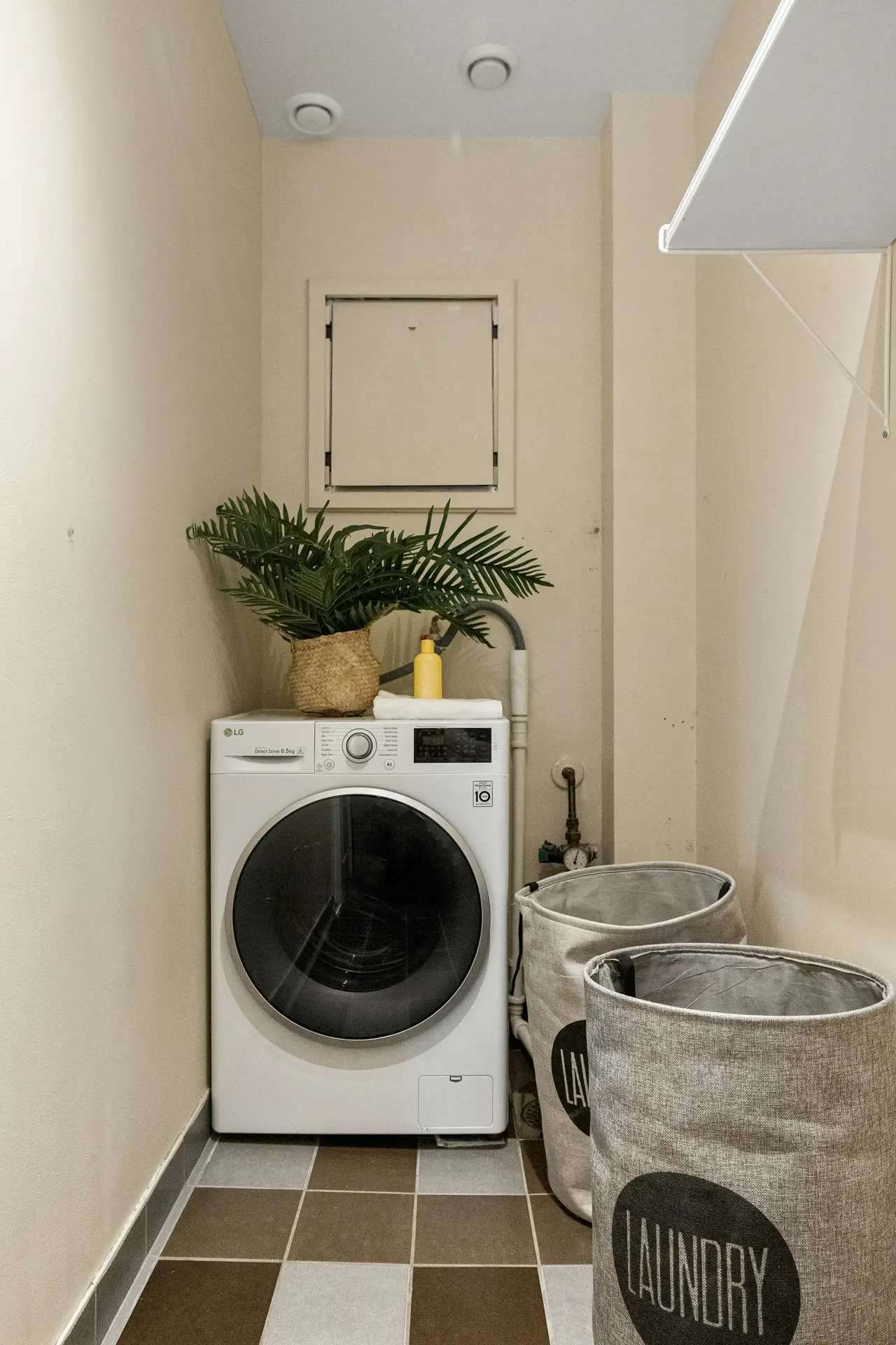A narrow laundry nook with a front-loading washing machine topped by a potted plant, neatly placed detergent, and two fabric laundry bags on the floor, showcasing laundry room organization ideas that keep small utility spaces tidy, functional, and visually calm.