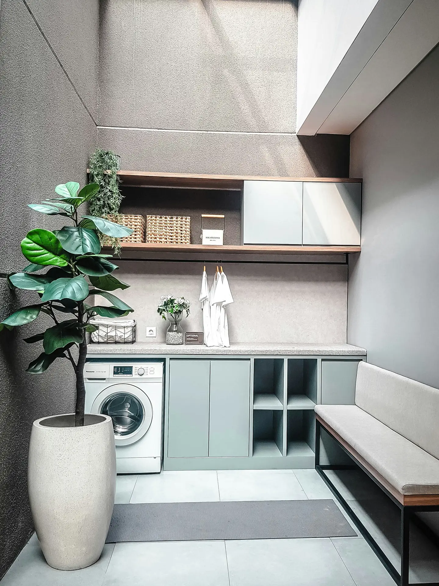 A modern, compact laundry space with built-in cabinets, open shelving, and a front-loading washer beneath a long countertop, paired with woven baskets, greenery, and a cushioned bench, showcasing laundry room organization ideas and a multi-functional laundry room that combines washing, storage, and seating in one streamlined layout.