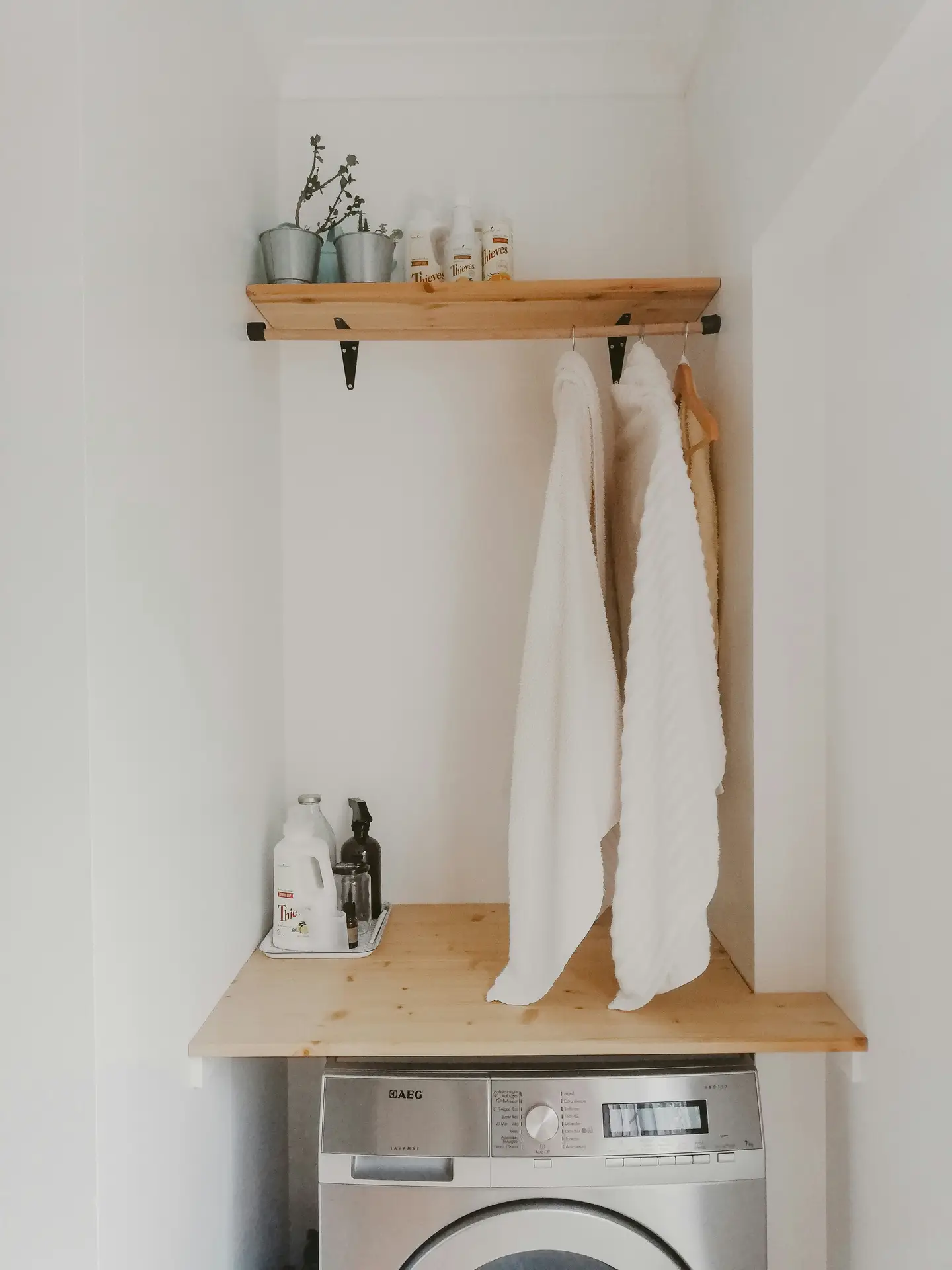 A compact laundry nook with a front-loading washing machine tucked beneath a wooden countertop, a shelf above laundry corner holding plants and cleaning products, and hooks with hanging towels, illustrating laundry room organization ideas that blend storage, practicality, and simple decor in a small space.