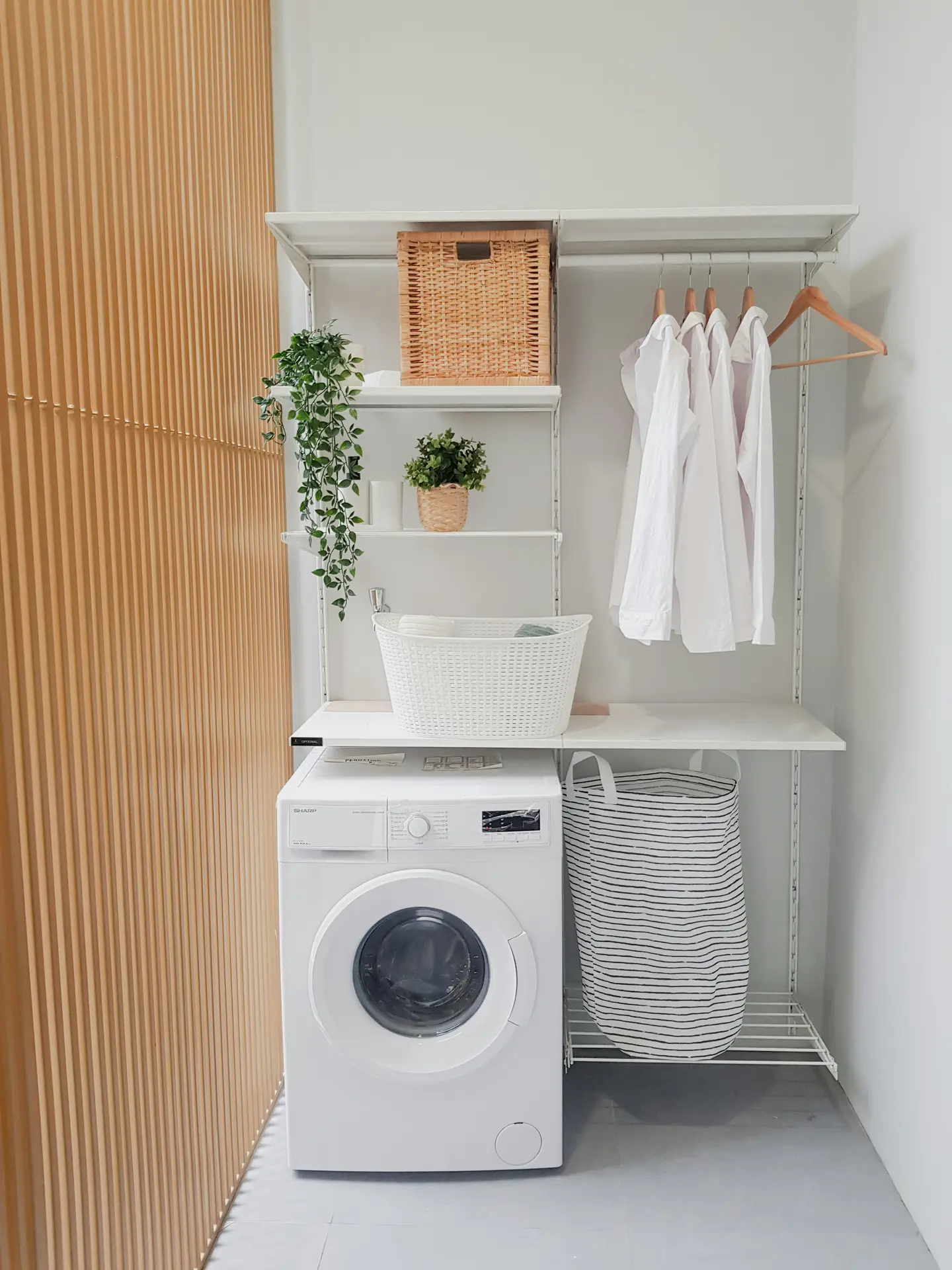 A compact laundry nook with a front-loading washing machine tucked beneath open shelving, featuring woven baskets, a hanging rail with neatly arranged shirts, potted plants, and a fabric laundry hamper, highlighting laundry room organization ideas and a smart laundry corner that blends storage, washing, and clothing care in one efficient setup.