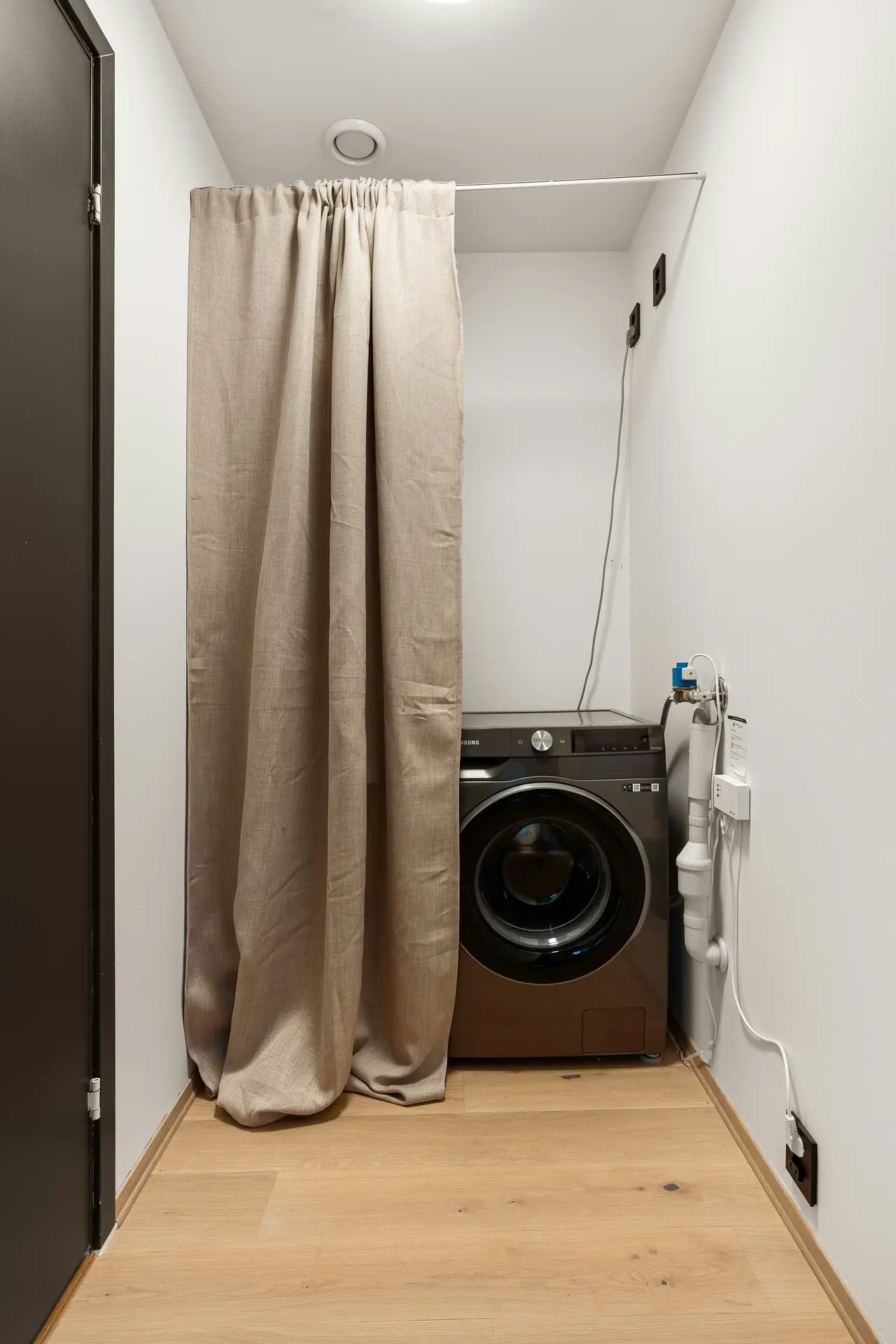 A narrow laundry alcove with a front-loading washing machine discreetly tucked behind a floor-length fabric curtain, showing laundry room organization ideas that use a washing mashine behind curtain to keep utilities hidden, maintain a clean look, and make the space feel calm and multifunctional.