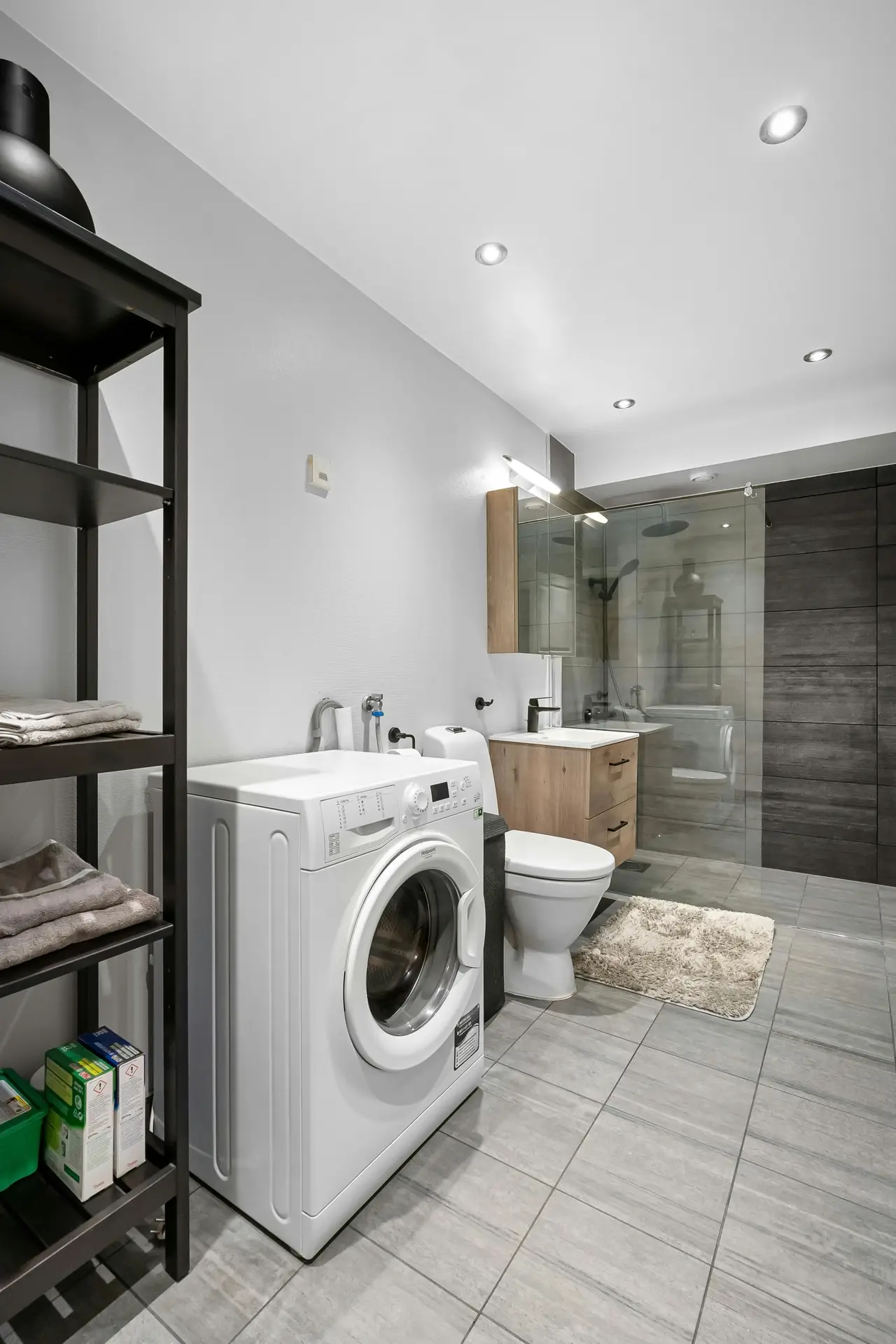 A modern bathroom featuring a front-loading washing machine placed beside a toilet and vanity, paired with open shelving for towels and detergents, showing laundry room organization ideas that integrate a washing machine in bathroom layouts without sacrificing style or functionality.
