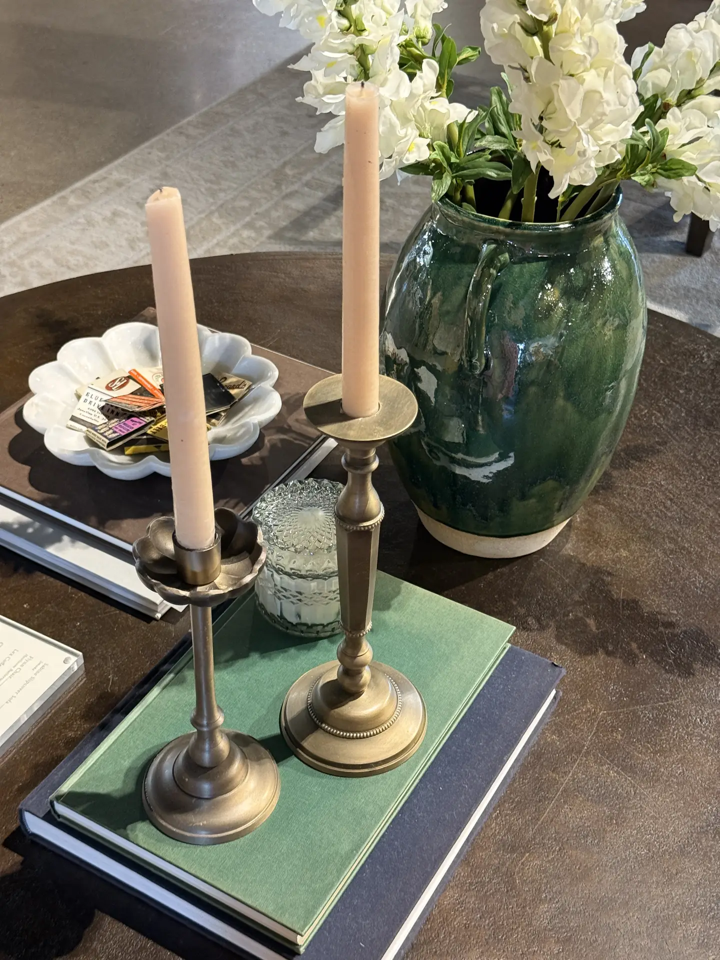 Living room coffee table decor ideas for spring showcasing candle holders on books, styled with two slim taper candles in antique-style metal holders placed on stacked hardcover books, paired with a textured glass jar, a small decorative dish, and a deep green ceramic vase filled with fresh white flowers, creating a layered and relaxed seasonal arrangement on a dark wooden coffee table.