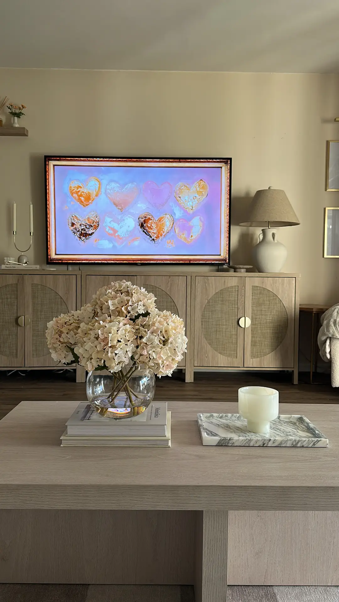 Living room coffee table decor ideas for spring featuring coffee table books with hydrangeas and marble tray with candle, styled in a calm neutral space with a low wooden table, a round glass vase filled with soft white hydrangeas, stacked books for height, and a simple marble tray holding a single candle, set against a cozy living room with a TV console, table lamps, and warm beige tones.
