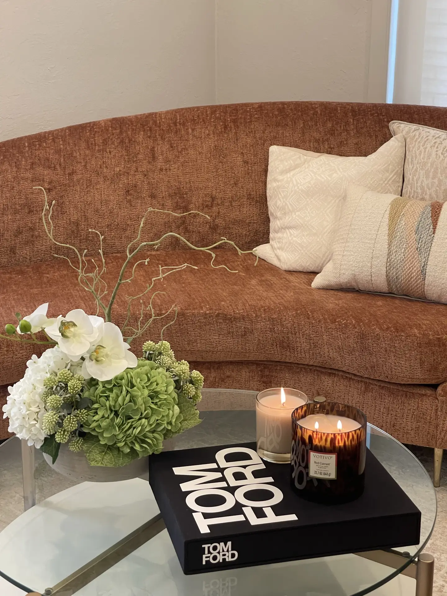Living room coffee table decor ideas for spring featuring flowers and coffee table book with candles, showing a glass coffee table styled with a soft green and white floral arrangement, a large black design book, and lit candles that add warmth in front of a curved rust-colored sofa with textured cushions.