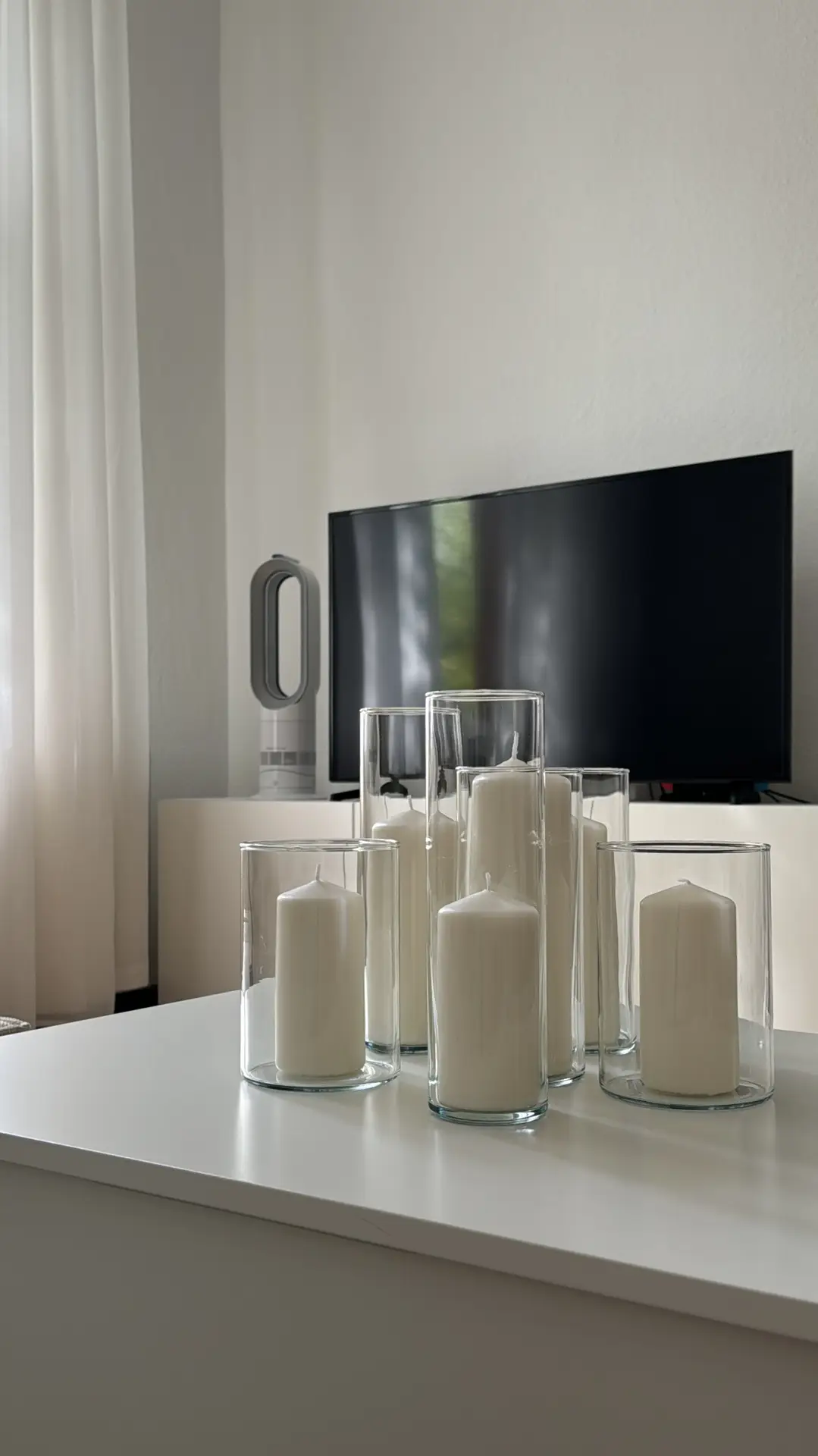 Living room coffee table decor ideas for spring featuring many white candles arranged in clear glass cylinders on a glossy white surface, creating a calm and airy look with soft daylight, sheer curtains, and a minimalist living room setting in the background.