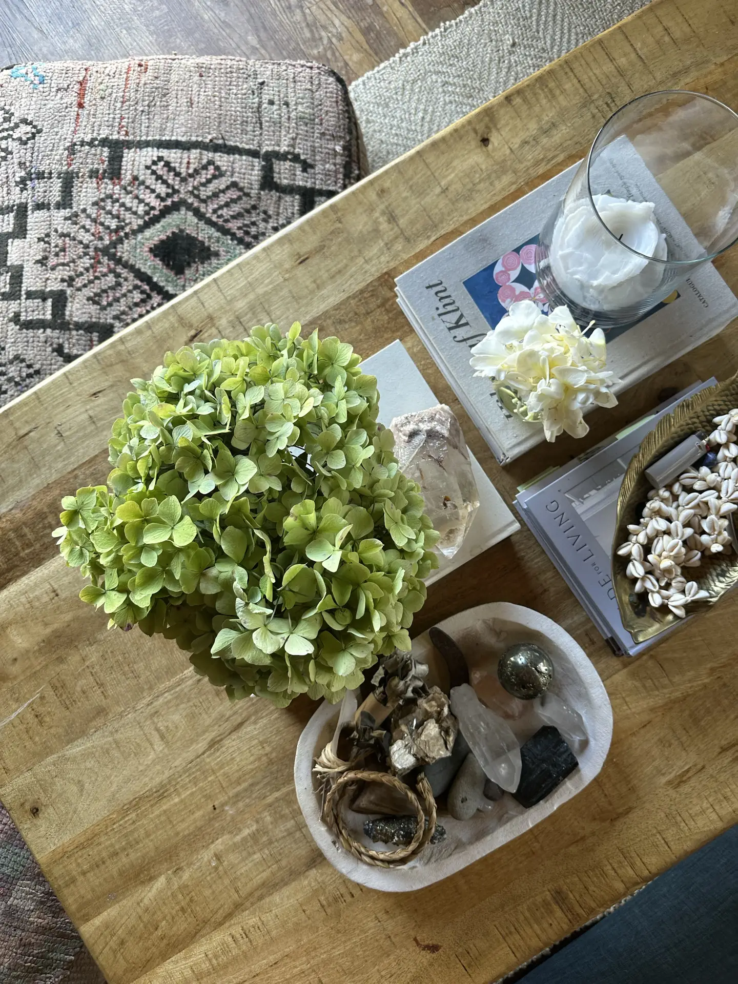 Living room coffee table decor ideas for spring with multiple decor accents, showing a wooden table styled with a soft green hydrangea arrangement, stacked books, a glass candle, shells, crystals, and natural objects arranged in small trays for a layered and relaxed look.