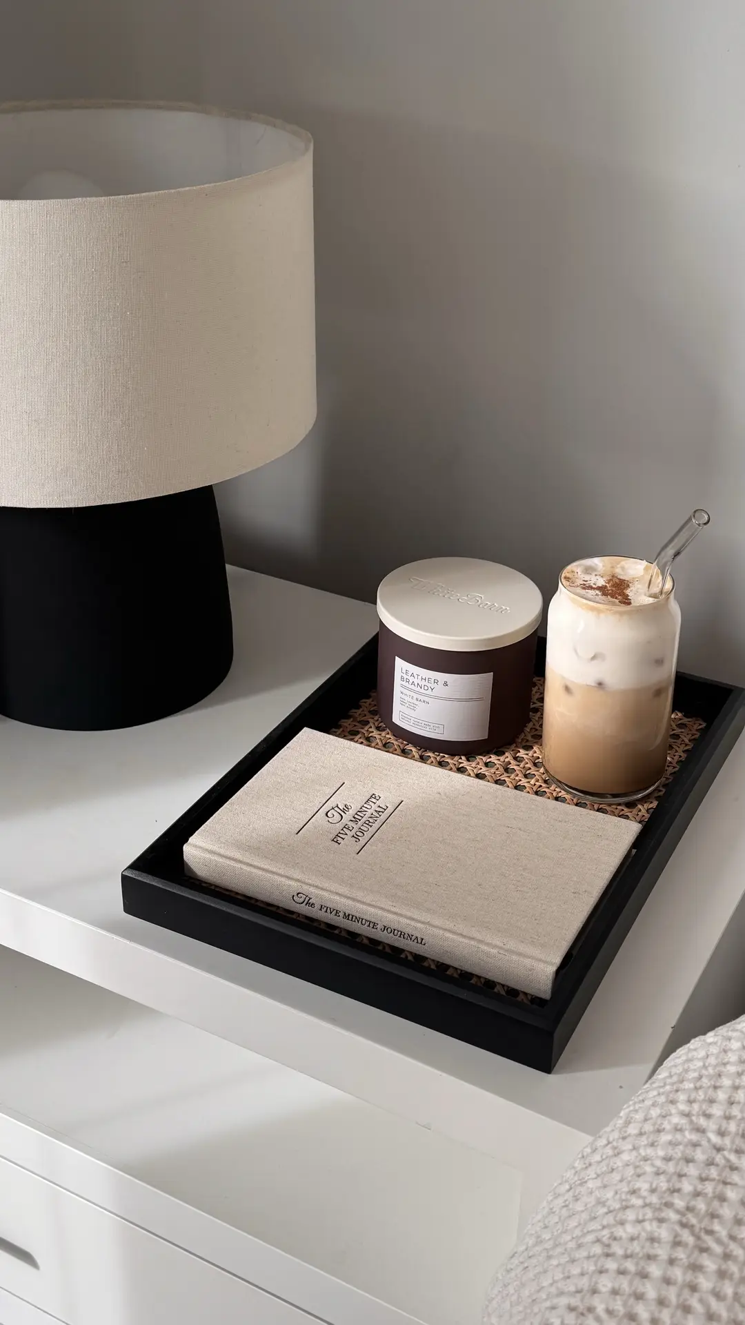 Living room coffee table decor ideas for spring showcasing a rattan tray styled with a neutral fabric-covered book, a scented candle, and an iced coffee glass, placed beside a modern table lamp, creating a relaxed and adaptable setup that fits both cozy and contemporary living spaces.