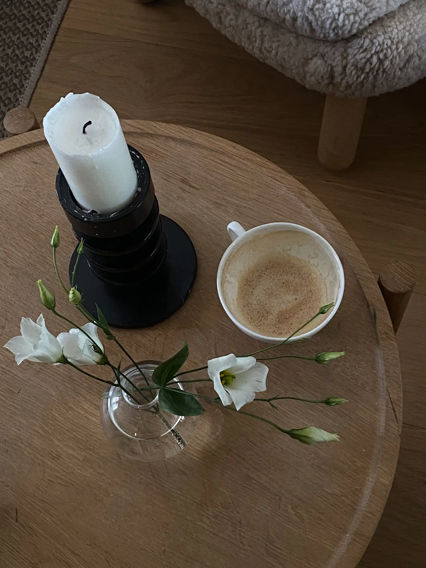 Living room coffee table decor ideas for spring featuring a white candle and minimalist white flowers, shown on a round wooden table styled with a black candle holder holding a white pillar candle, a small glass vase with delicate white blooms and buds, and a cup of coffee, creating a calm, natural, and cozy atmosphere.