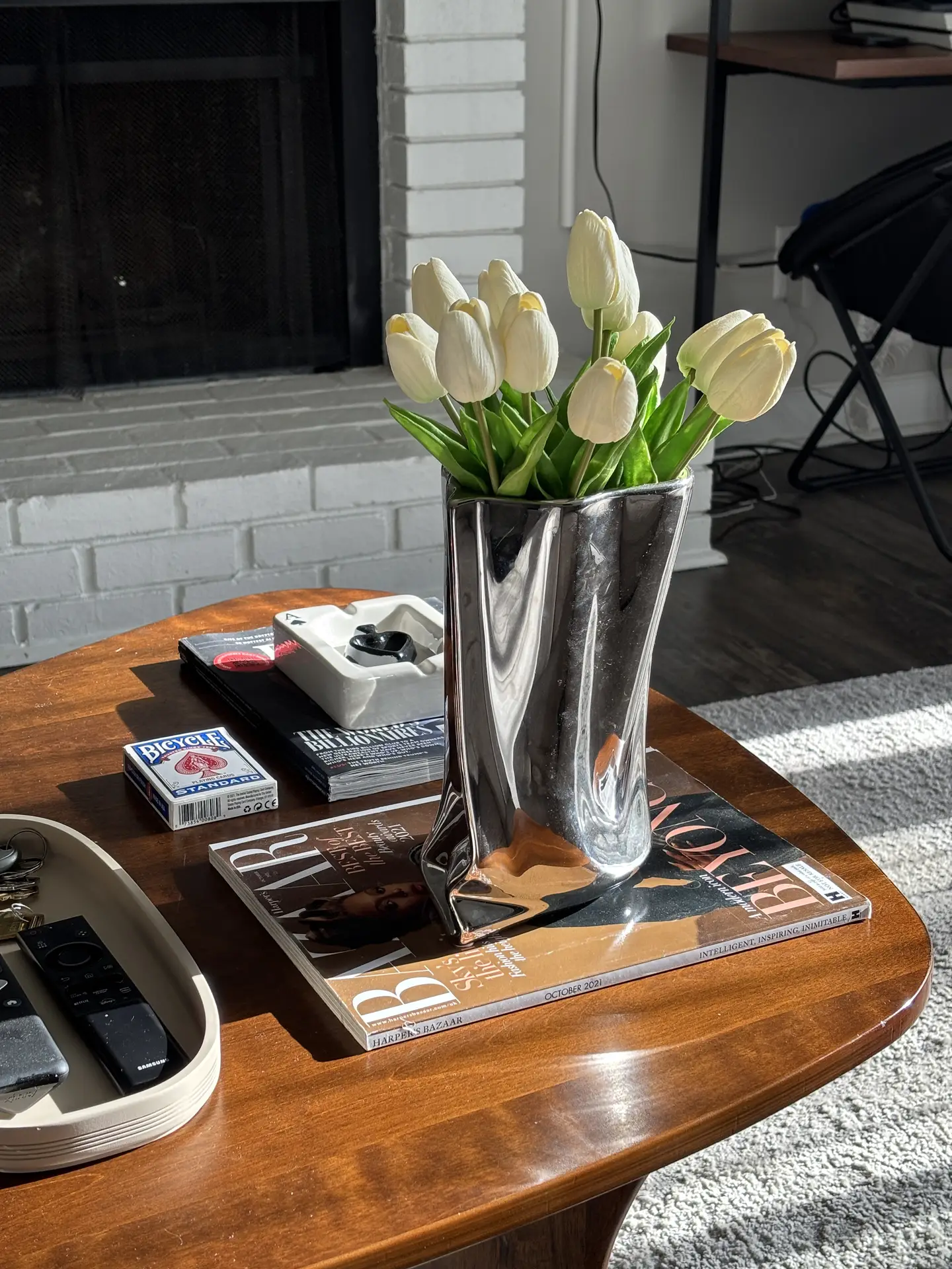 Living room coffee table decor ideas for spring featuring white tulips on magazine, showing a warm wooden coffee table styled with a sculptural glass vase holding fresh white tulips placed on stacked fashion magazines, alongside small everyday items, all lit by natural sunlight in a cozy living room setting.