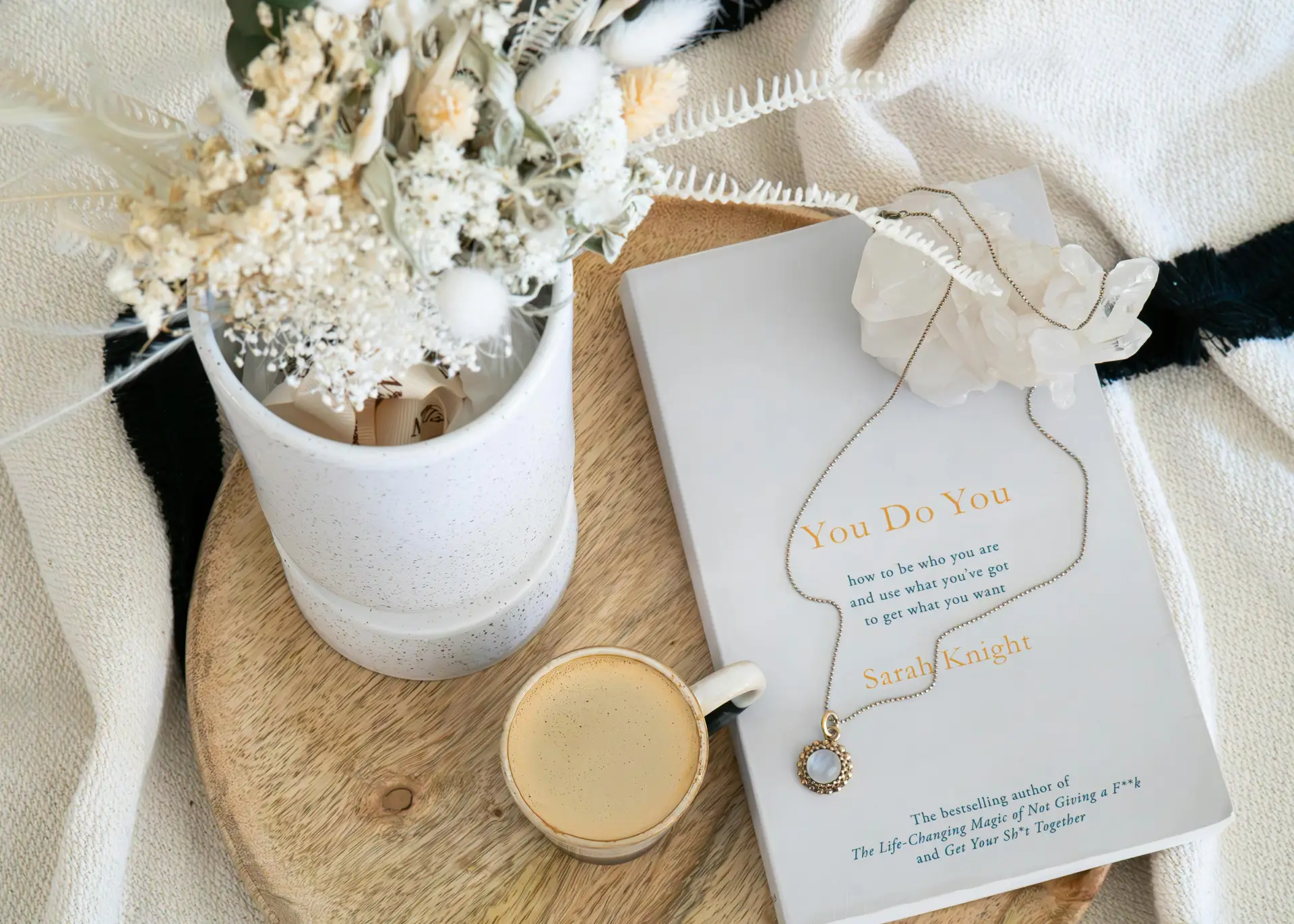 Living room coffee table decor ideas for spring featuring a wooden tray with dried flowers arranged in a neutral vase, a matching soft-toned book with a delicate necklace placed on top, and a small cup of coffee, creating a calm, layered look that works well for cozy, minimalist, or modern living spaces.