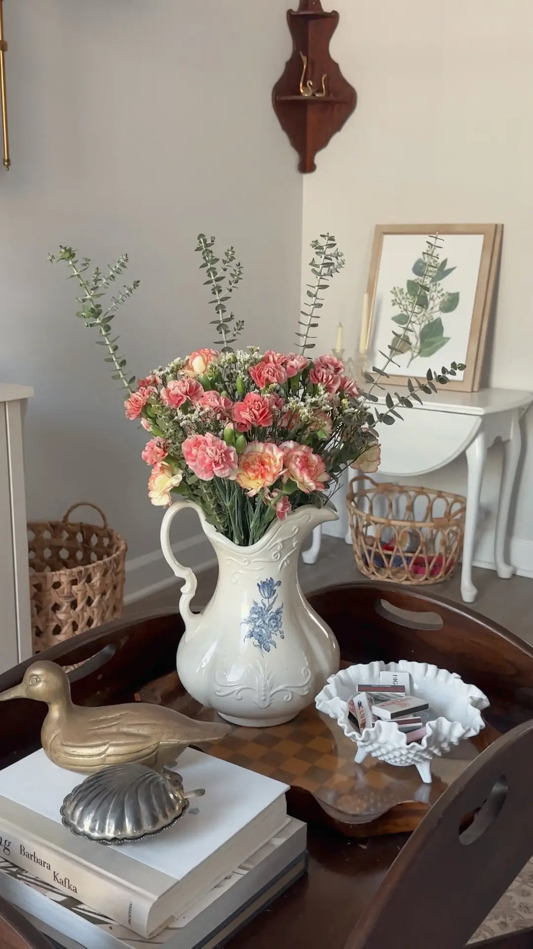 Living room coffee table decor ideas for spring styled with a wooden tray with flowers and decor, featuring a vintage-style ceramic pitcher filled with soft pink blooms and eucalyptus, layered books topped with a small brass bird accent, and a sculptural white bowl, all arranged to create a warm, curated look with classic and natural elements.