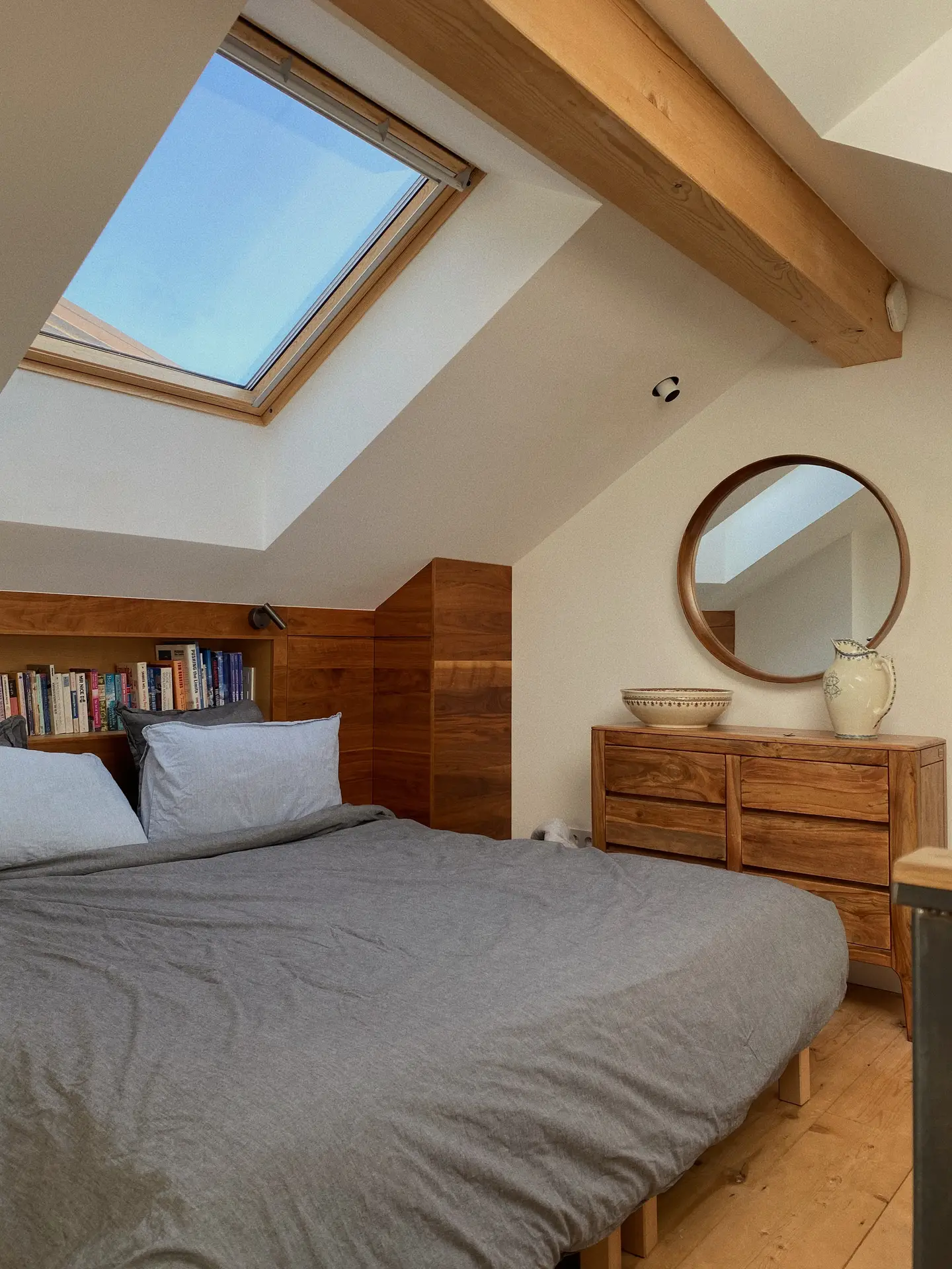 A cozy attic bedroom with a skylight features a wooden bed frame that includes built-in shelves behind the pillows, filled with books, alongside a matching dresser and simple decor, illustrating organization ideas for small spaces by using headboard storage to combine sleeping and storage in one streamlined setup.