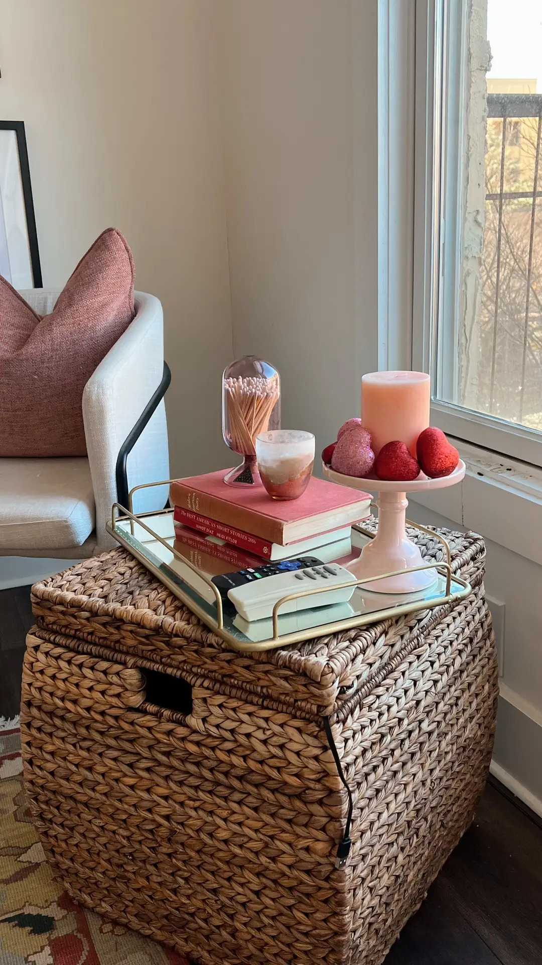 A woven rattan cube placed beside a sofa is styled as a small side table with a tray holding books, candles, and a remote, showcasing organization ideas for small spaces by using a multi-functional stool that works as seating, storage, and a decorative surface near a window.