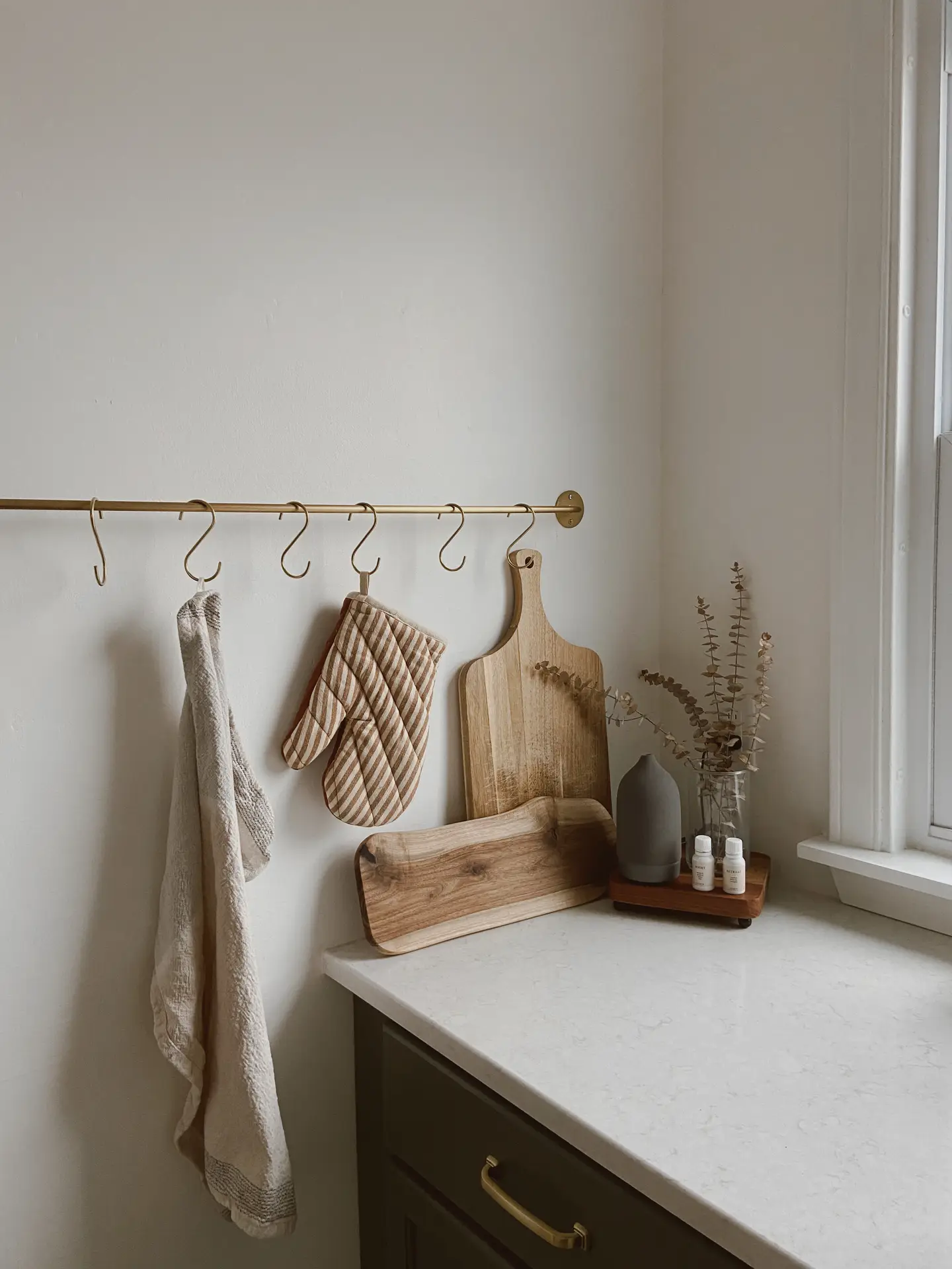 Minimal kitchen corner showing organization ideas for small spaces, featuring a slim rail with hooks holding a towel and oven mitt, wooden cutting boards leaning against the wall, and a tidy countertop styled with neutral decor near a window.