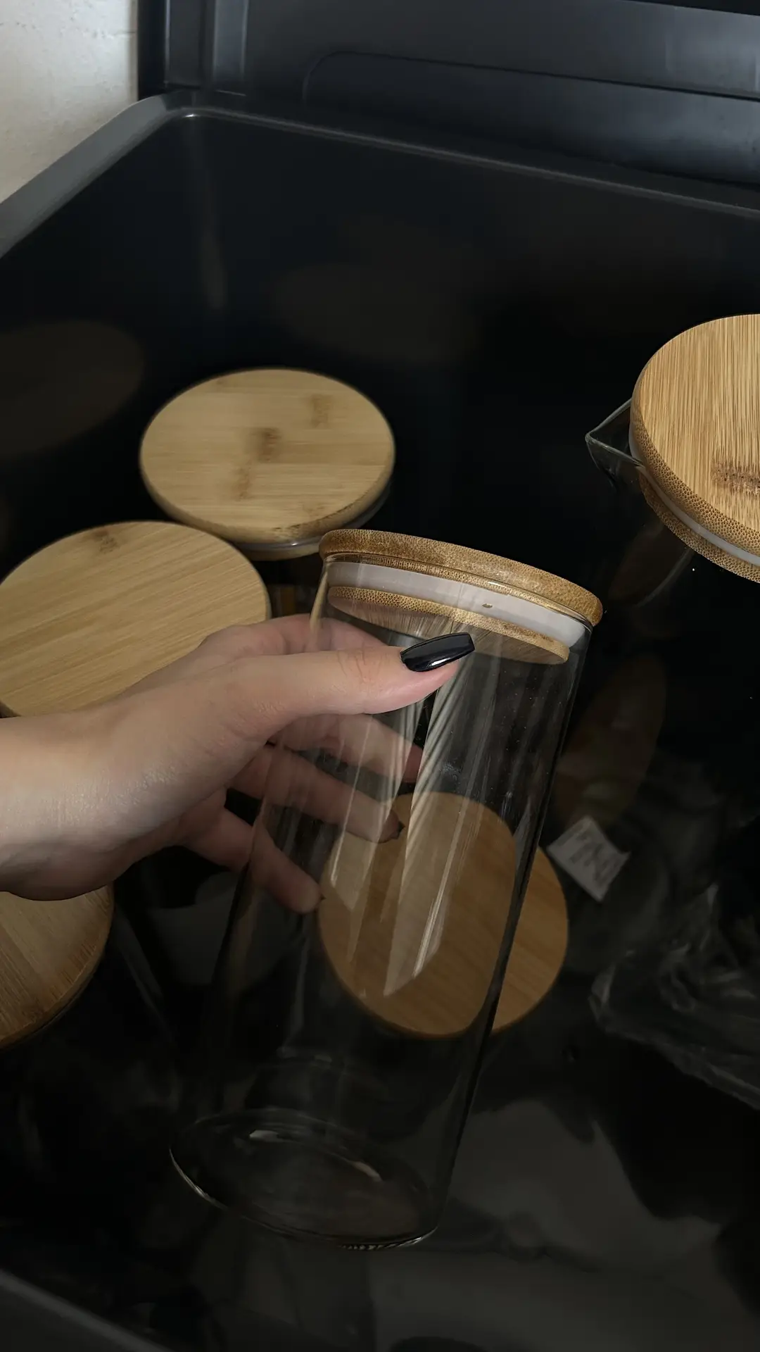 Close-up of a hand holding a clear glass jar with a bamboo lid inside a drawer, surrounded by matching containers, illustrating organization ideas for small spaces where you use glas containers to keep storage tidy, uniform, and easy to access.