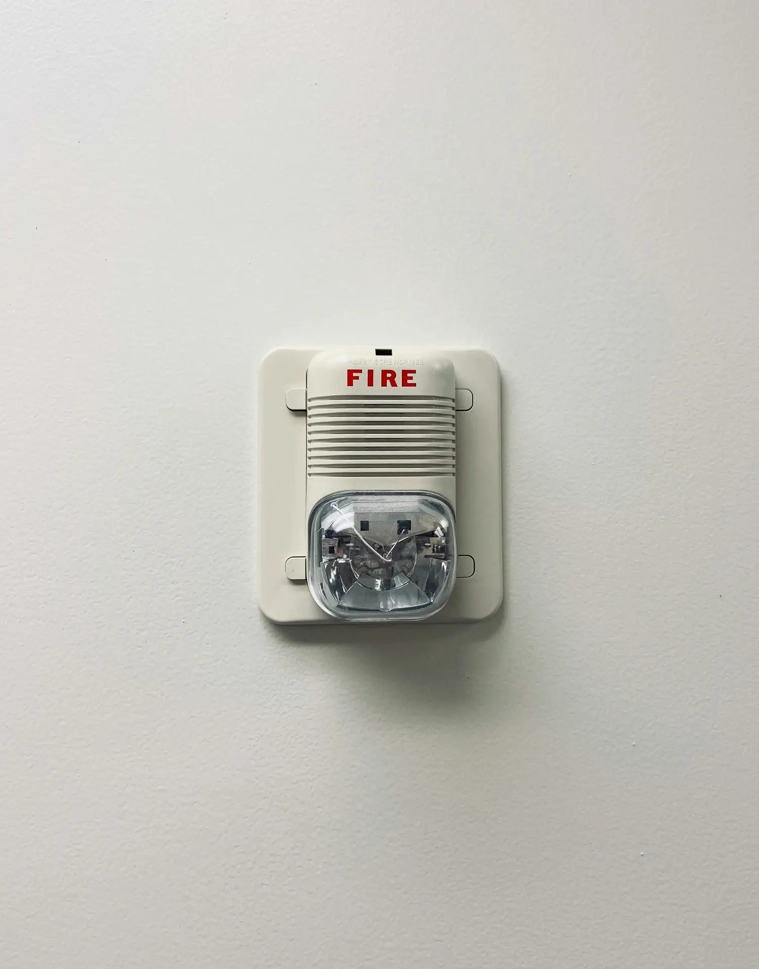 Wall-mounted fire alarm with a clear strobe light and red fire label on a plain white wall, fitting into a spring cleaning checklist reminder to check the batteries in smoke detectors as part of regular home safety maintenance.