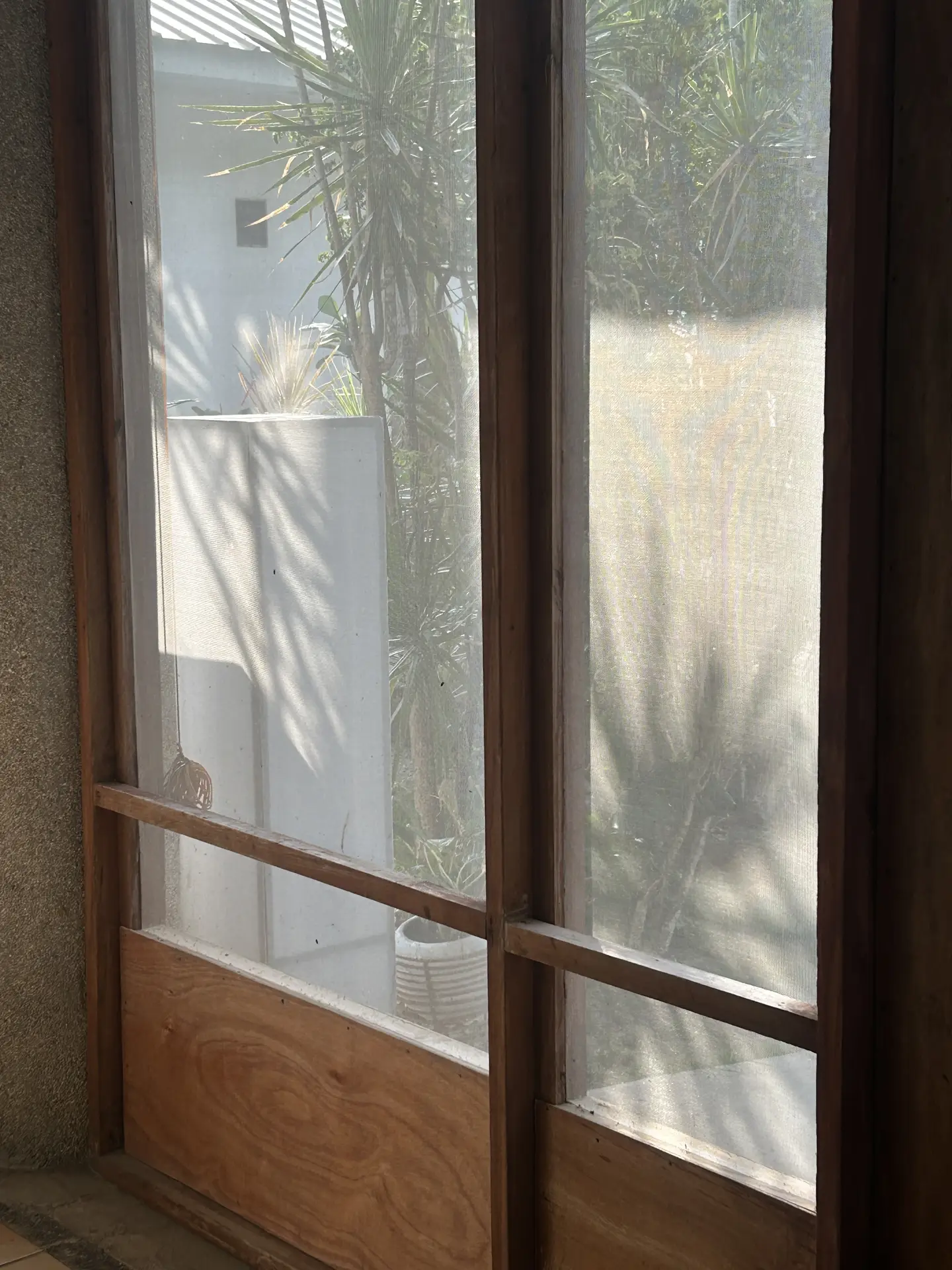 A sunlit window with visible mesh fly screens and a wooden frame, showing outdoor greenery behind slightly dusty screens, illustrating a spring cleaning checklist task focused on cleaning fly screens to improve airflow and natural light indoors.